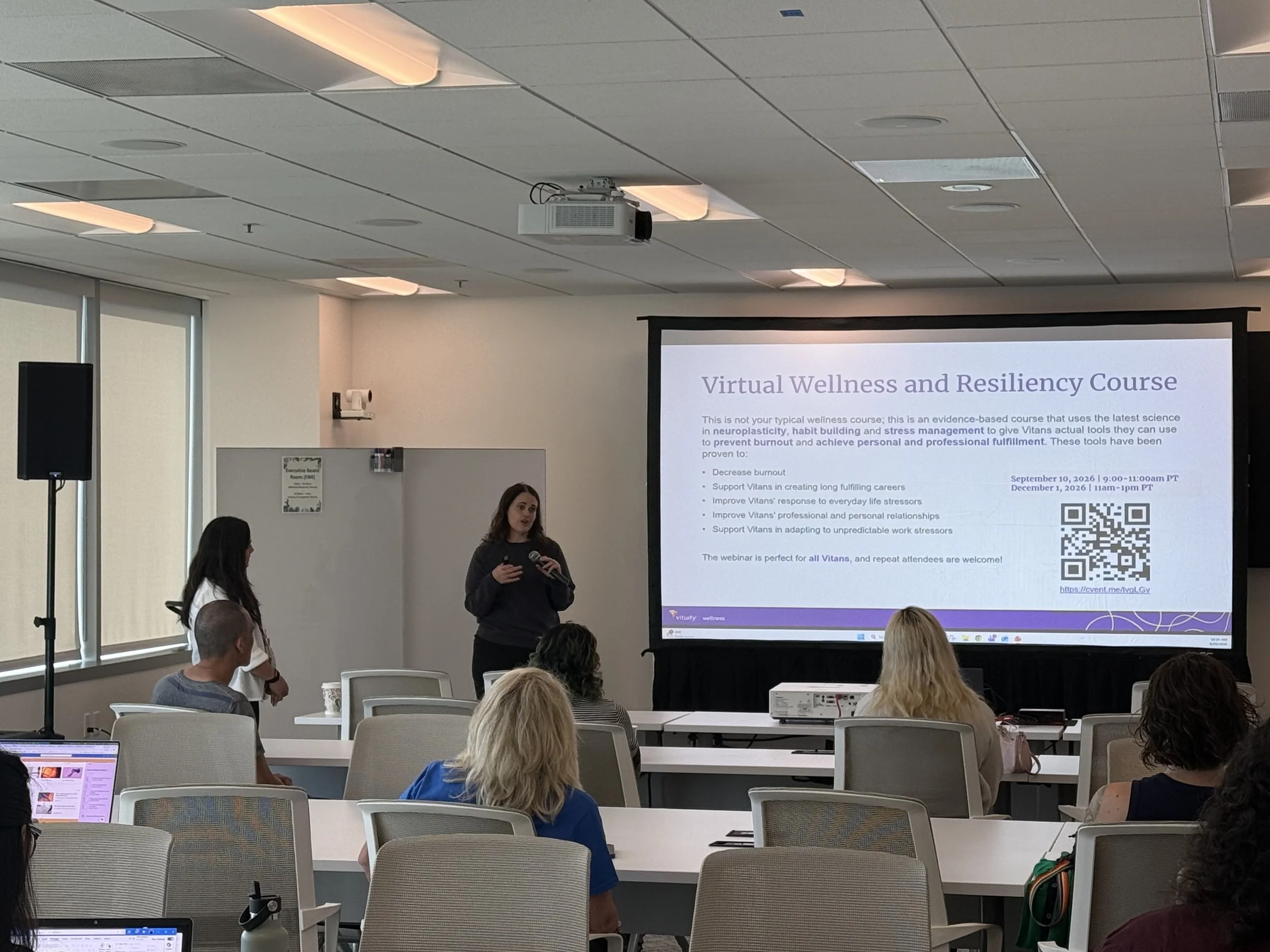 A woman is giving a presentation in a conference room titled 'Virtual Wellness and Resiliency Course.' Attendees are seated, some with laptops open, listening to the speaker. The room has ceiling lights, a projector, and a large presentation screen displaying course information.