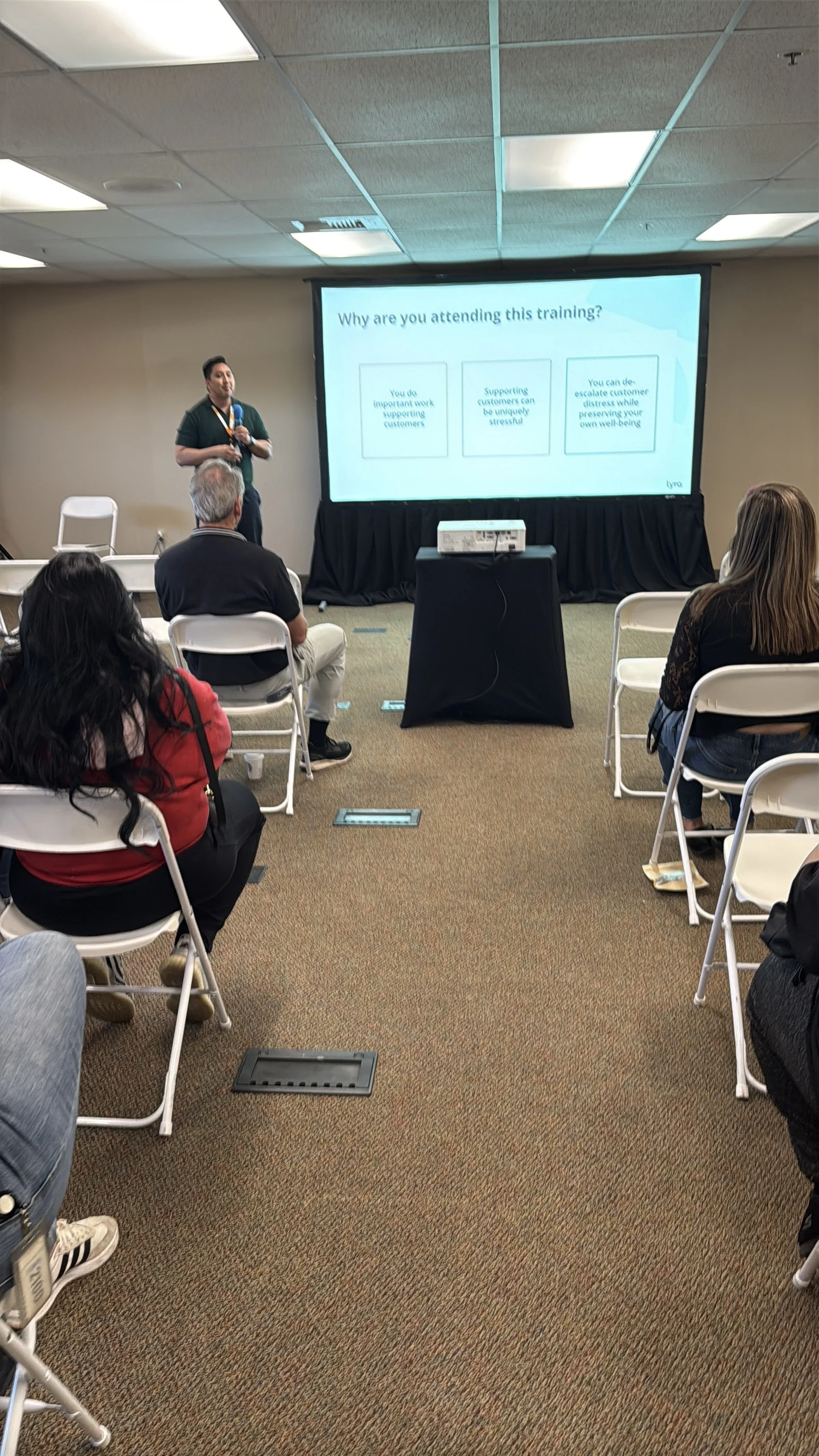 A person holding a microphone giving a presentation in a conference room with a projector screen displaying a slide titled 'Why are you attending this training?' with three bullet points.