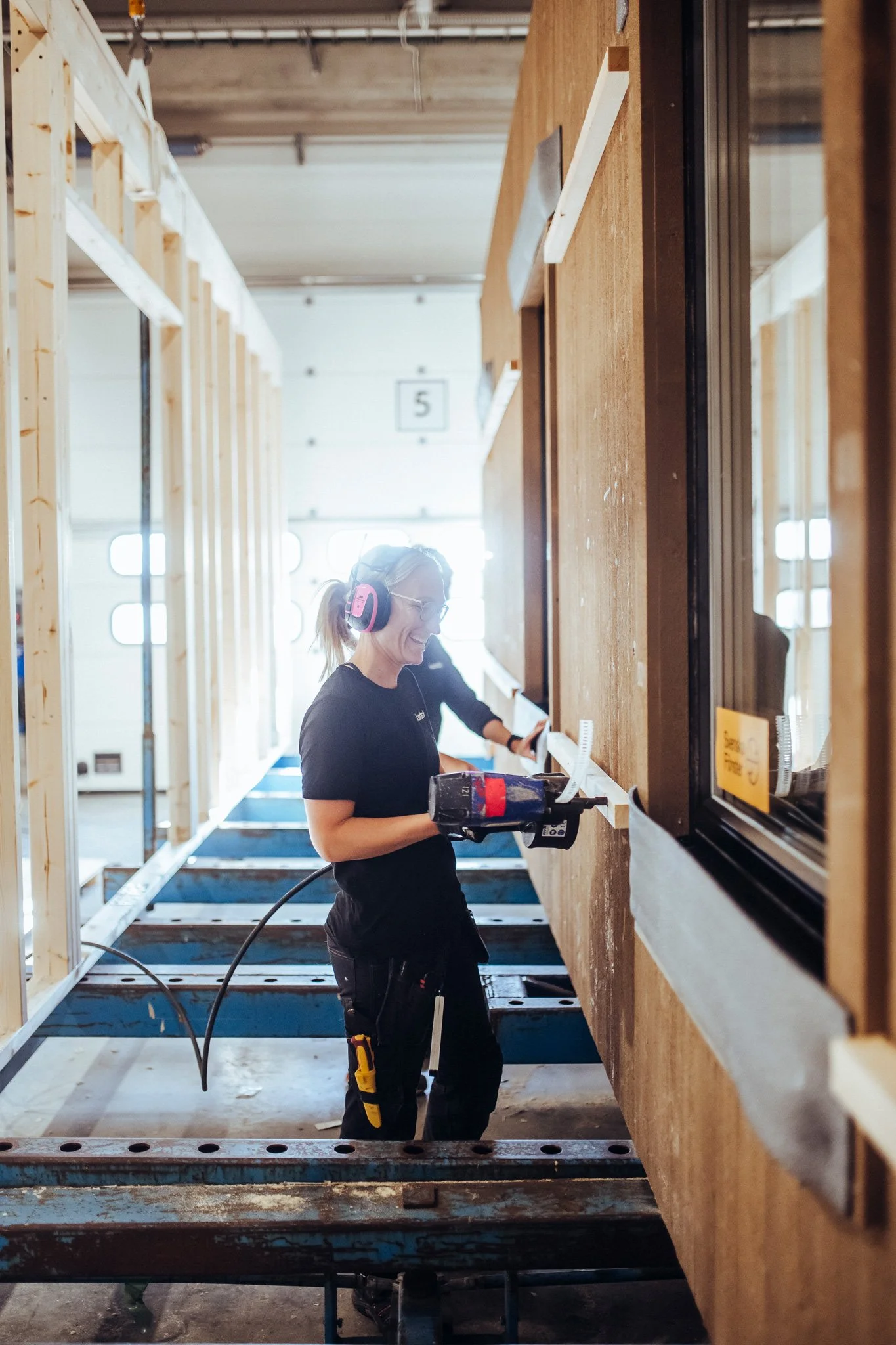 A woman wearing safety headphones and glasses using a power drill while working on a construction project inside a building with exposed wooden framing.