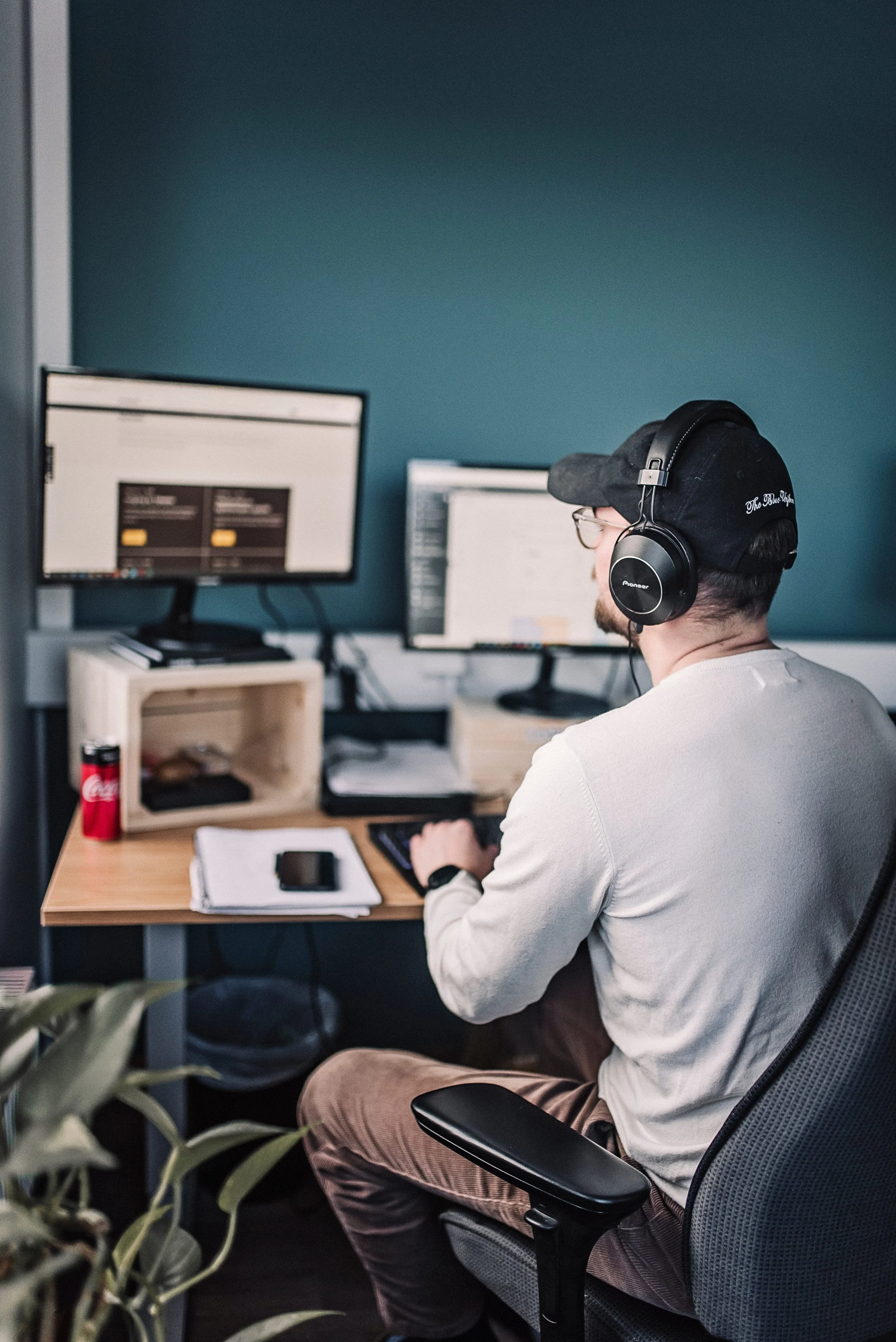 A man wearing a black cap, glasses, and headphones sitting at a desk working on a computer with two monitors in an office. There is a smartphone, a notebook, and a can of Coca-Cola on the desk.