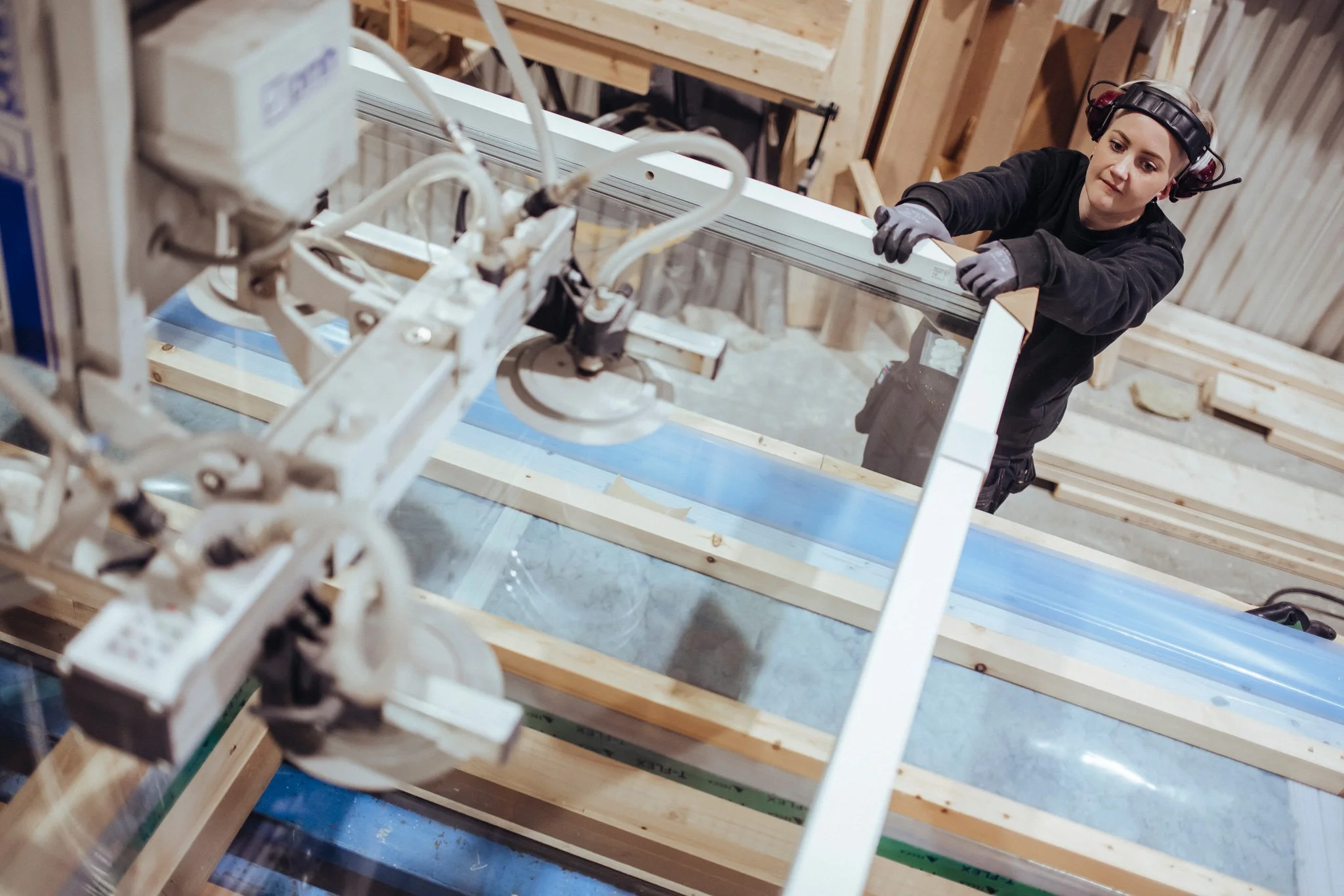 A woman working on a construction project in a workshop, wearing safety glasses and ear protection, handling a framed glass panel.