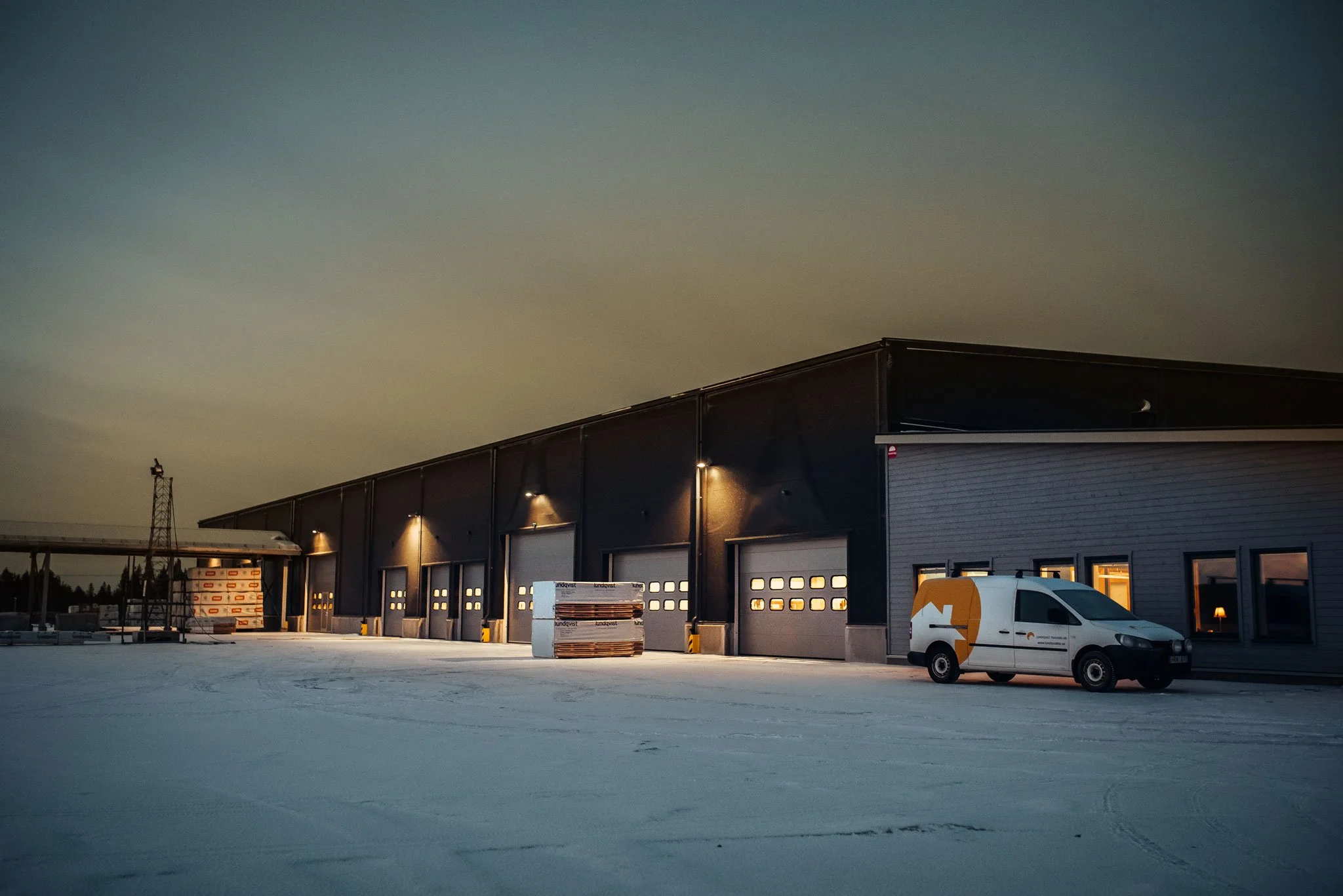 An industrial warehouse with multiple garage doors, outdoor lighting, and a white delivery van parked in front, under a darkening sky at dusk.