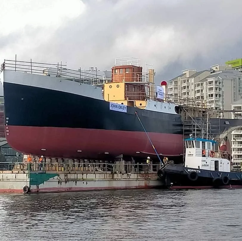 A large ship named 'John Oxley' is being transported on a barge along a river or canal in an urban area with high-rise buildings in the background. Several workers and a tugboat are present around the vessel.
