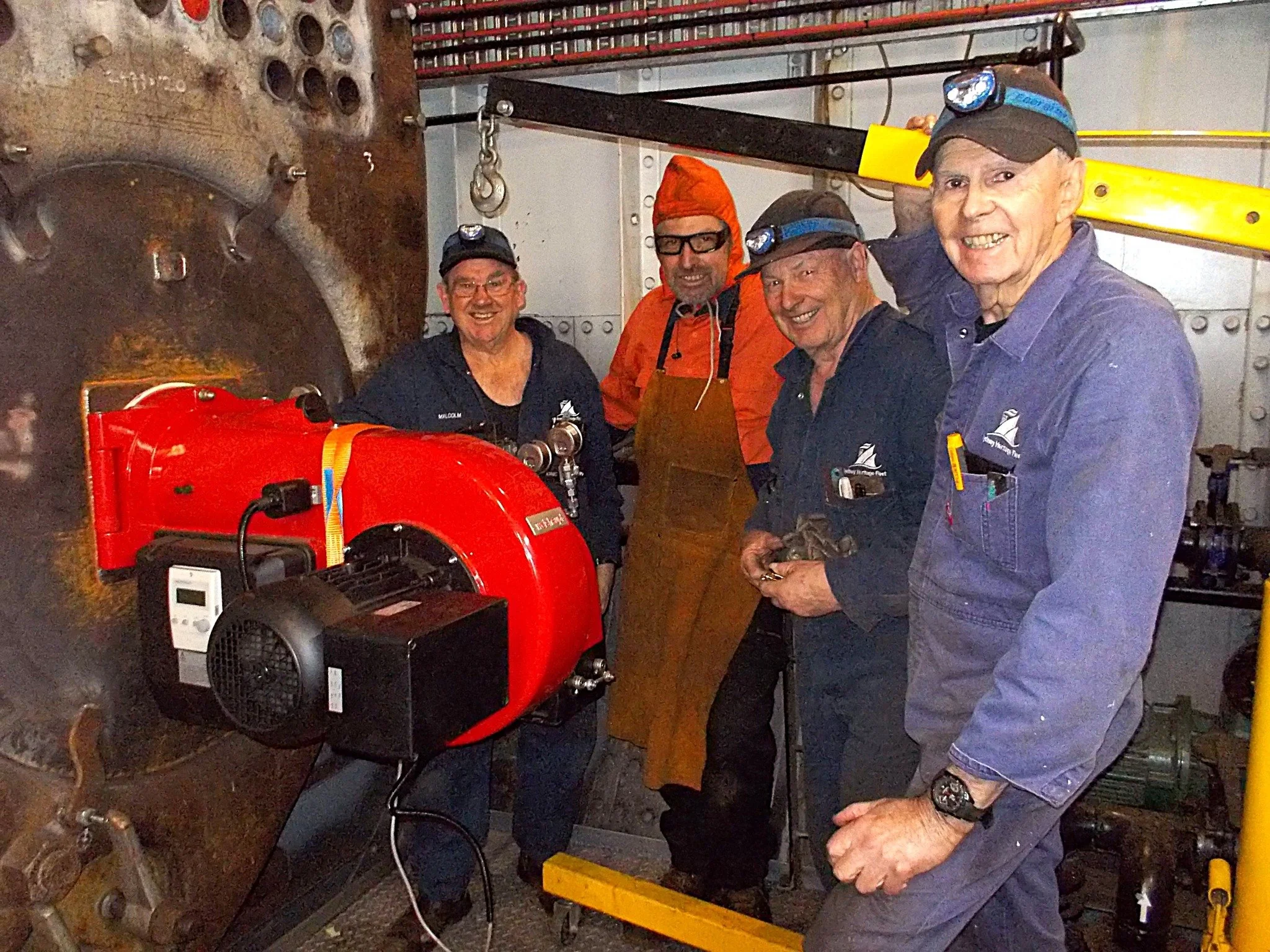 Group of five men working inside an industrial or ship engine room, with machinery and equipment around them.