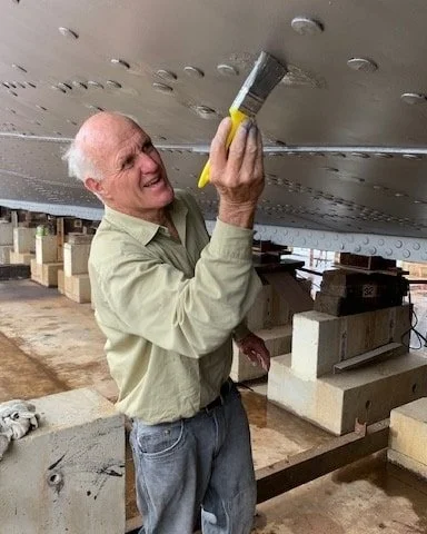 An elderly man in a light-colored shirt and jeans is smiling while painting the underside of a large boat with a brush.