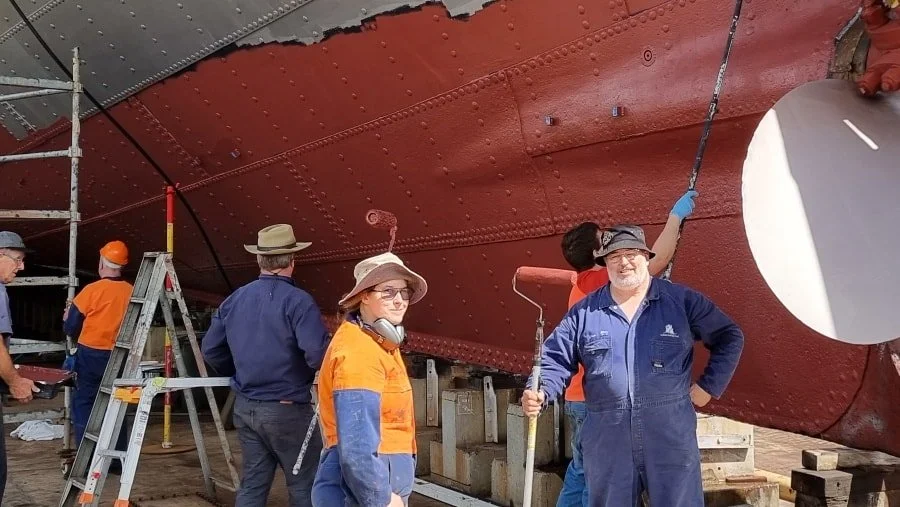 People working on the underside of a large ship hull, which is being painted red, with some on ladders and others standing on the ground.