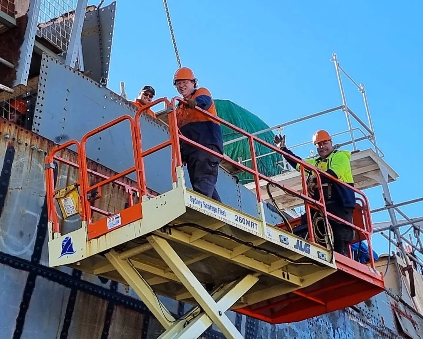 Construction workers wearing safety vests and helmets working on a building scaffold, standing on an orange aerial work platform against a blue sky.