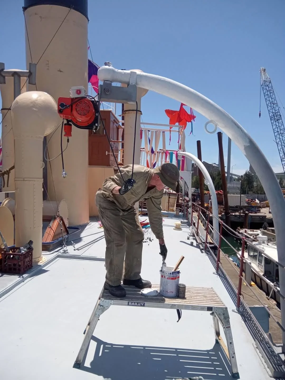 A man standing on a step ladder painting the deck of a ship, with construction equipment and flags visible in the background.