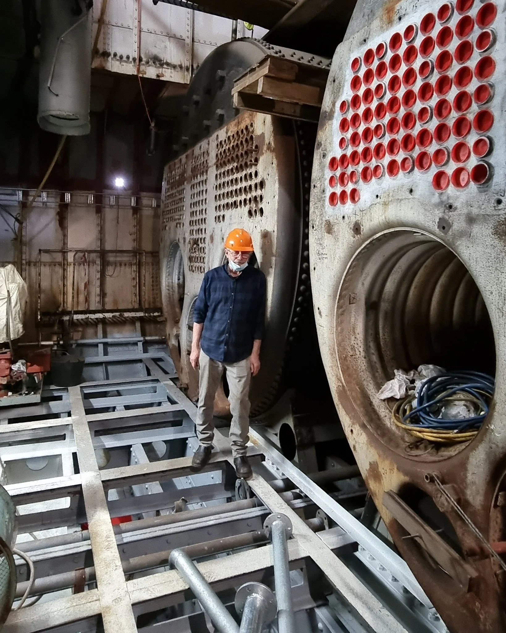 A man wearing a yellow safety helmet, safety glasses, and a face mask standing inside a large industrial machine or turbine with rusty and clean parts, on a metal scaffolding framework.