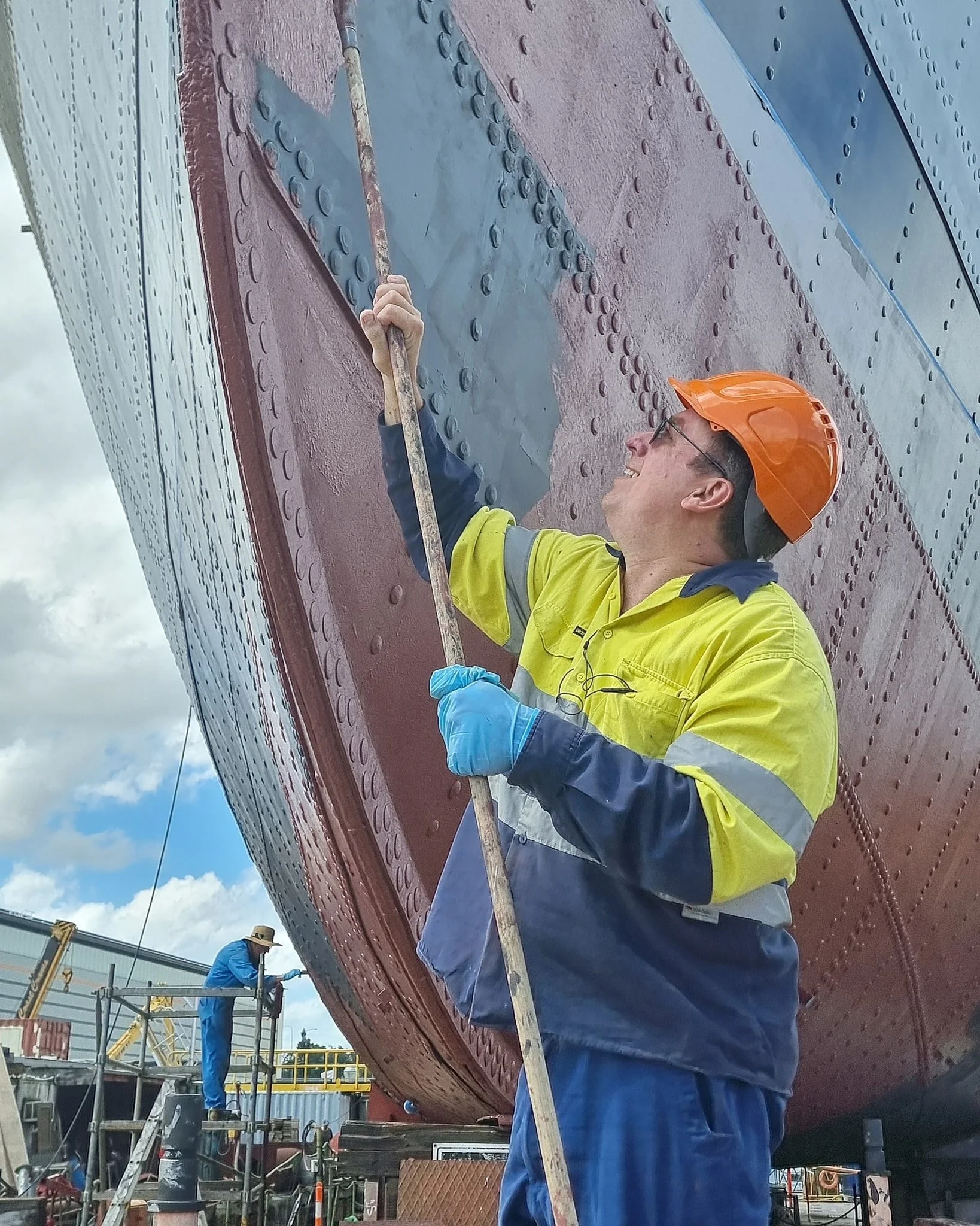 A worker wearing an orange helmet, glasses, a yellow and navy safety jacket, and blue gloves is using a long tool to work on a large, riveted metal ship hull.