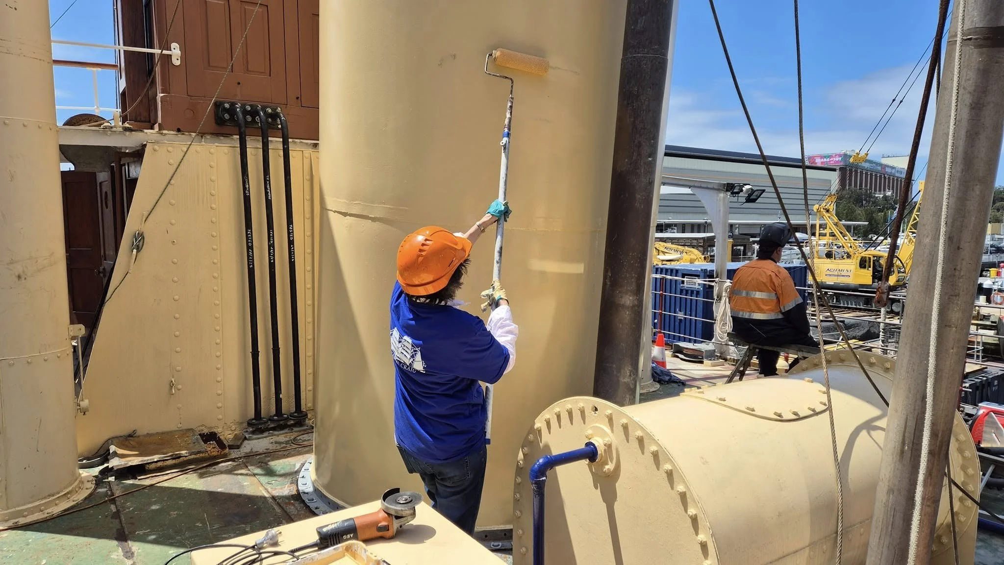 Worker in blue shirt and orange helmet using a roller brush on large yellow industrial equipment at a construction site.