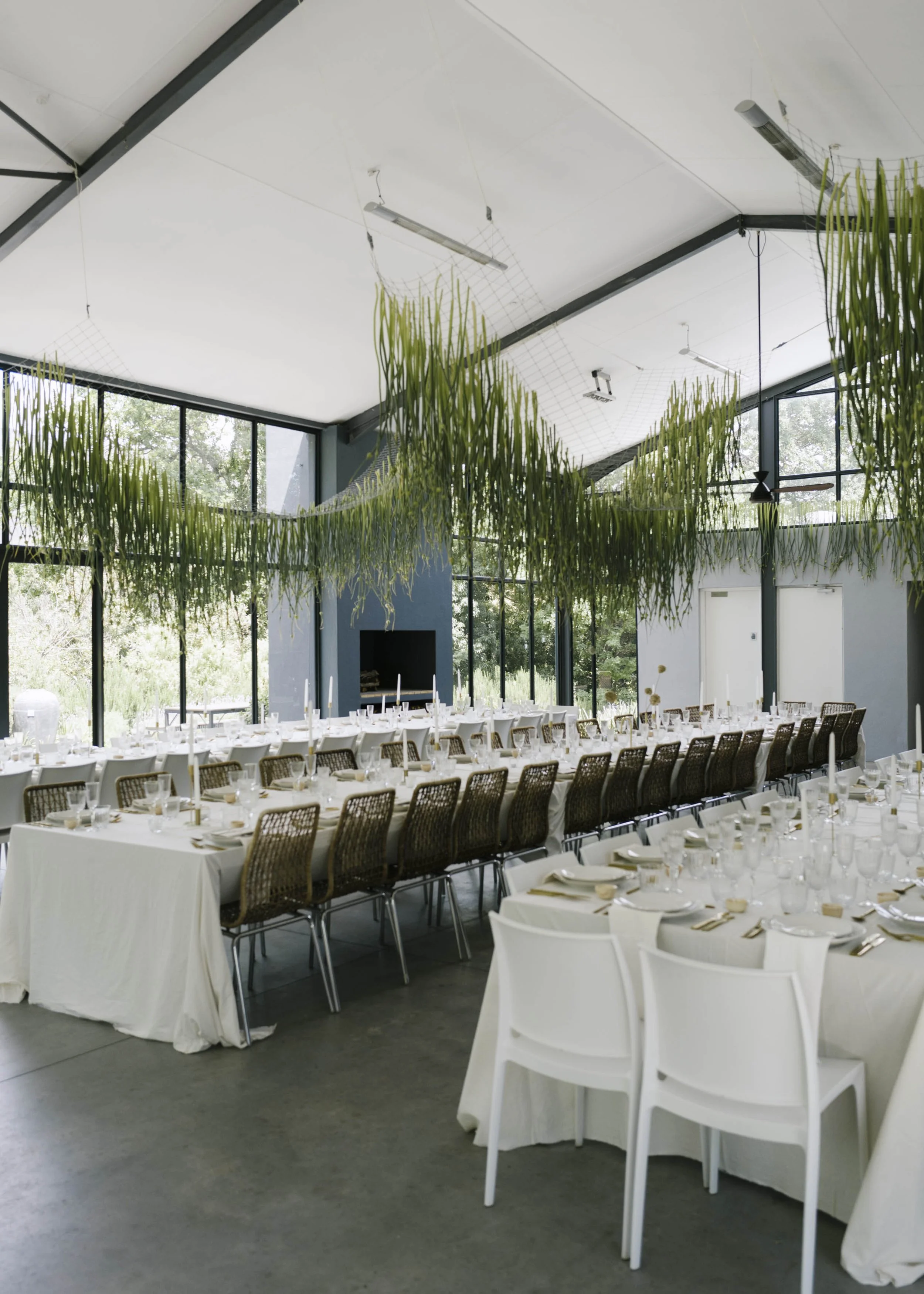 Interior of a large, bright event space with long tables set for a meal, white tablecloths, and white chairs. There are large windows with a view of greenery outside, and green plants hanging from the ceiling.