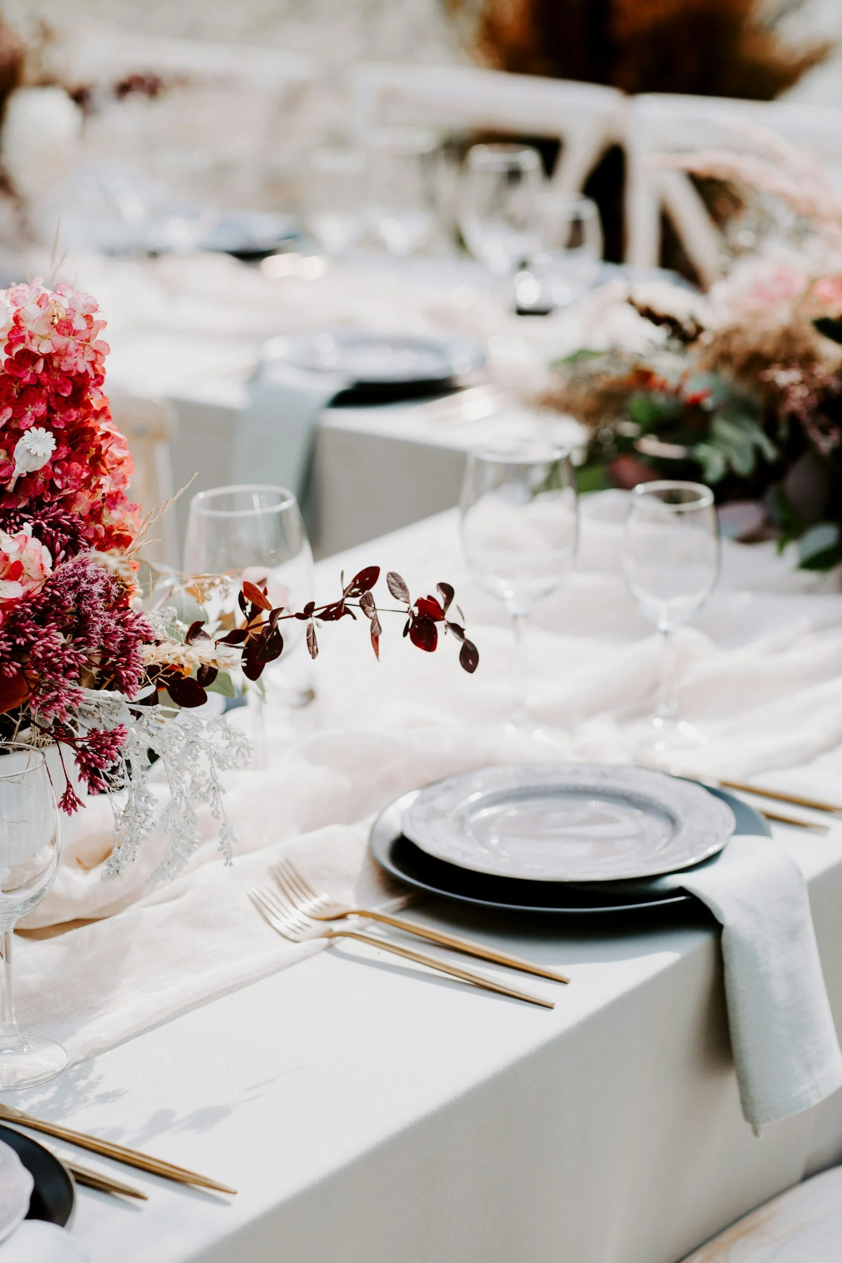 Elegant outdoor wedding table setting with floral centerpiece, white tablecloth, gold flatware, black and white plates, and clear wine glasses, decorated with greenery and soft natural light.