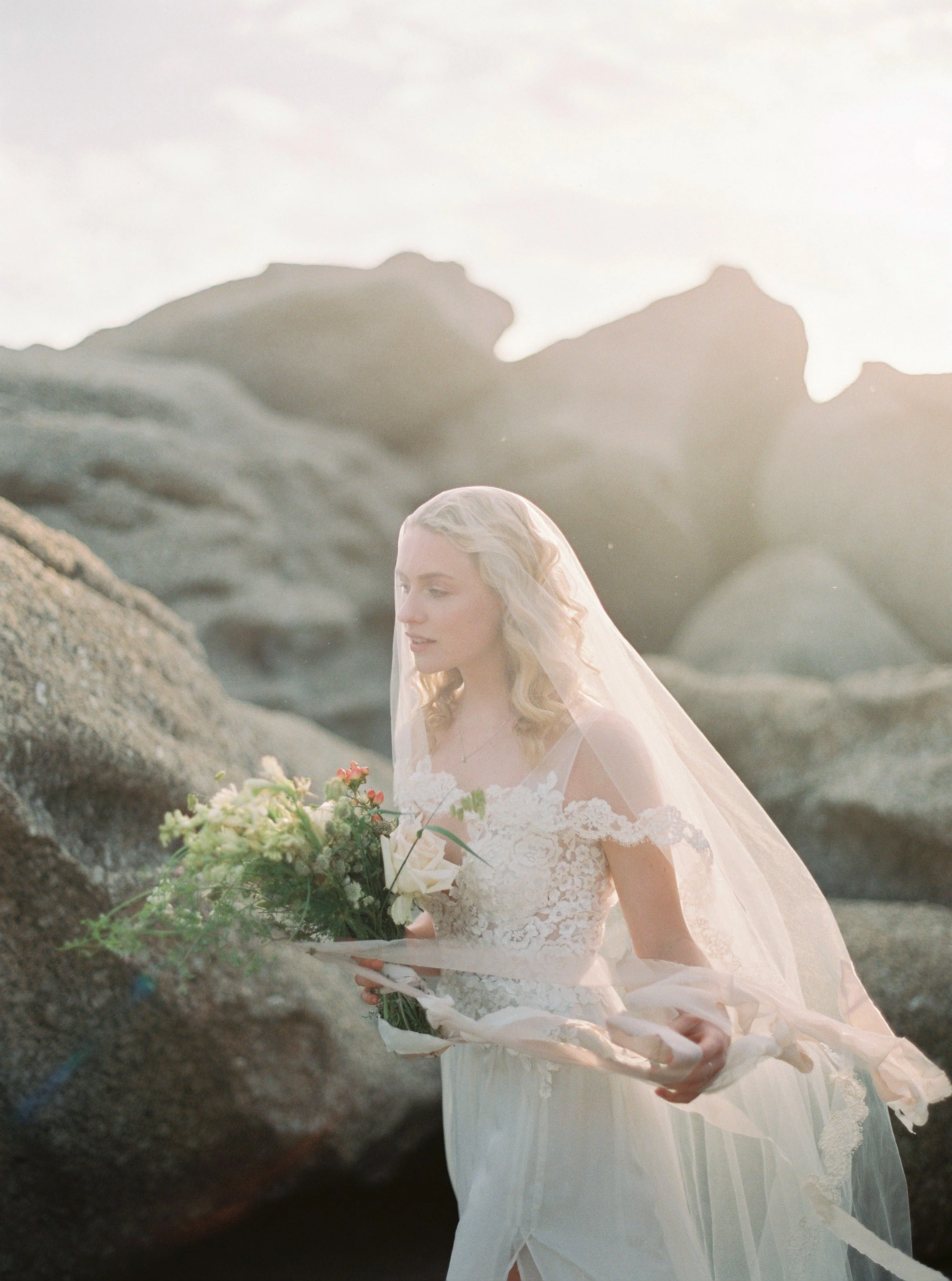 A bride in a white wedding dress and veil holds a bouquet of flowers on rocky terrain, with her head bowed, during sunset or sunrise.
