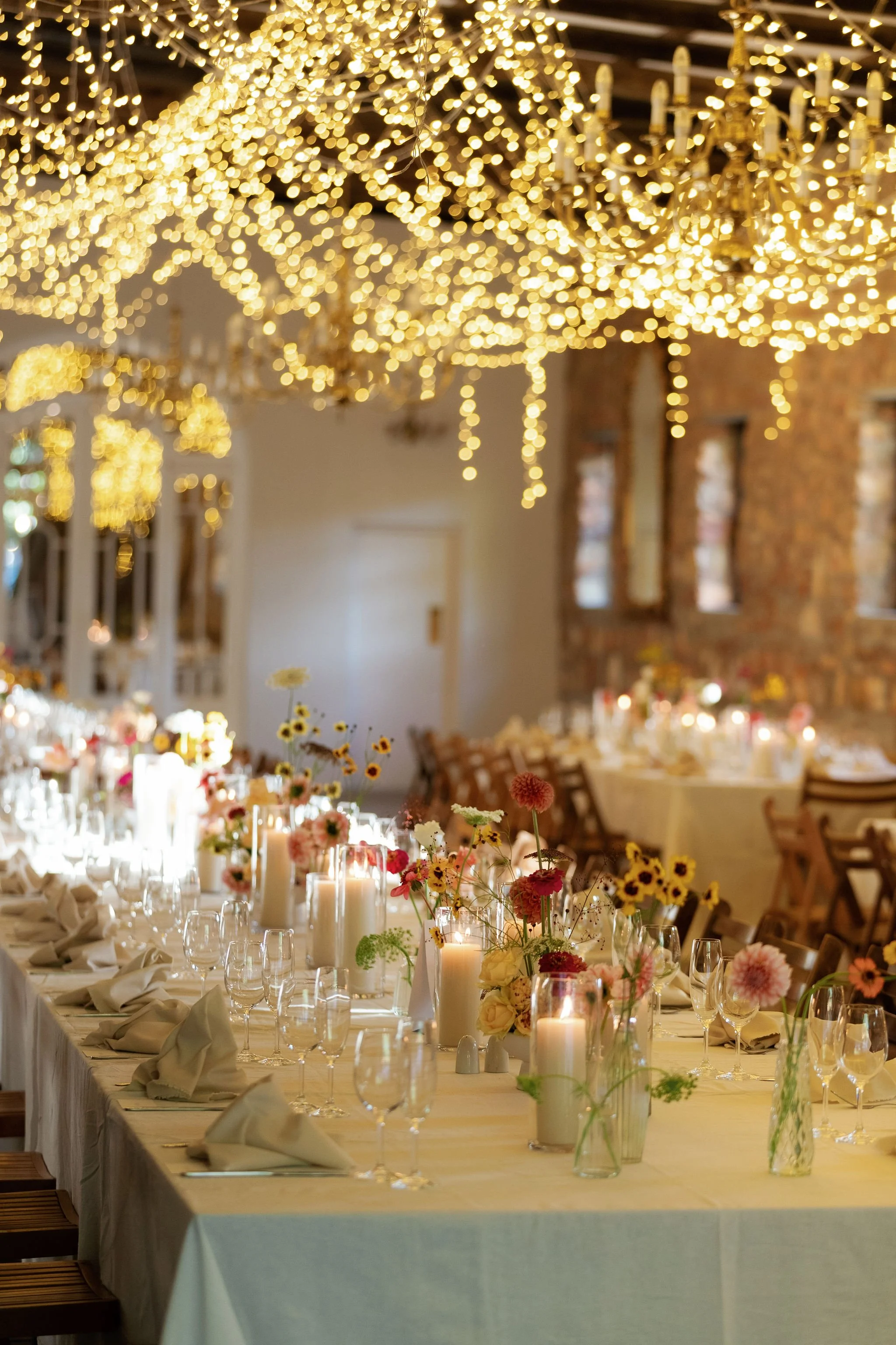 A decorated banquet hall with string lights and chandeliers, long tables set with flowers, candles, glasses, and folded napkins, ready for a celebration or event.