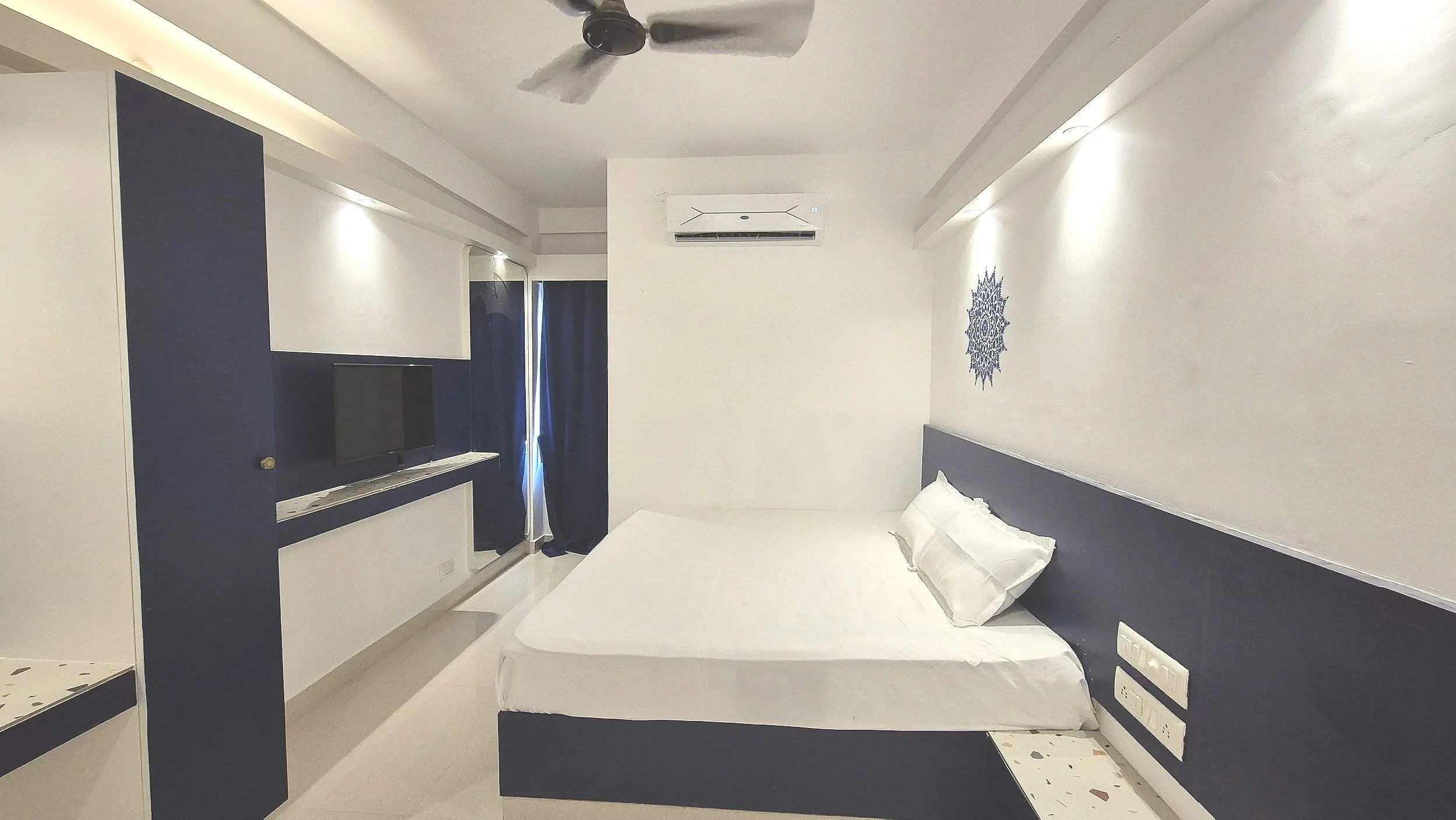 A modern, small hotel room with a white bed and white pillows, navy blue wall accents, a mounted flat-screen TV, a dark-colored desk or shelf, a ceiling fan, an air conditioning unit, and a window with navy blue curtains.