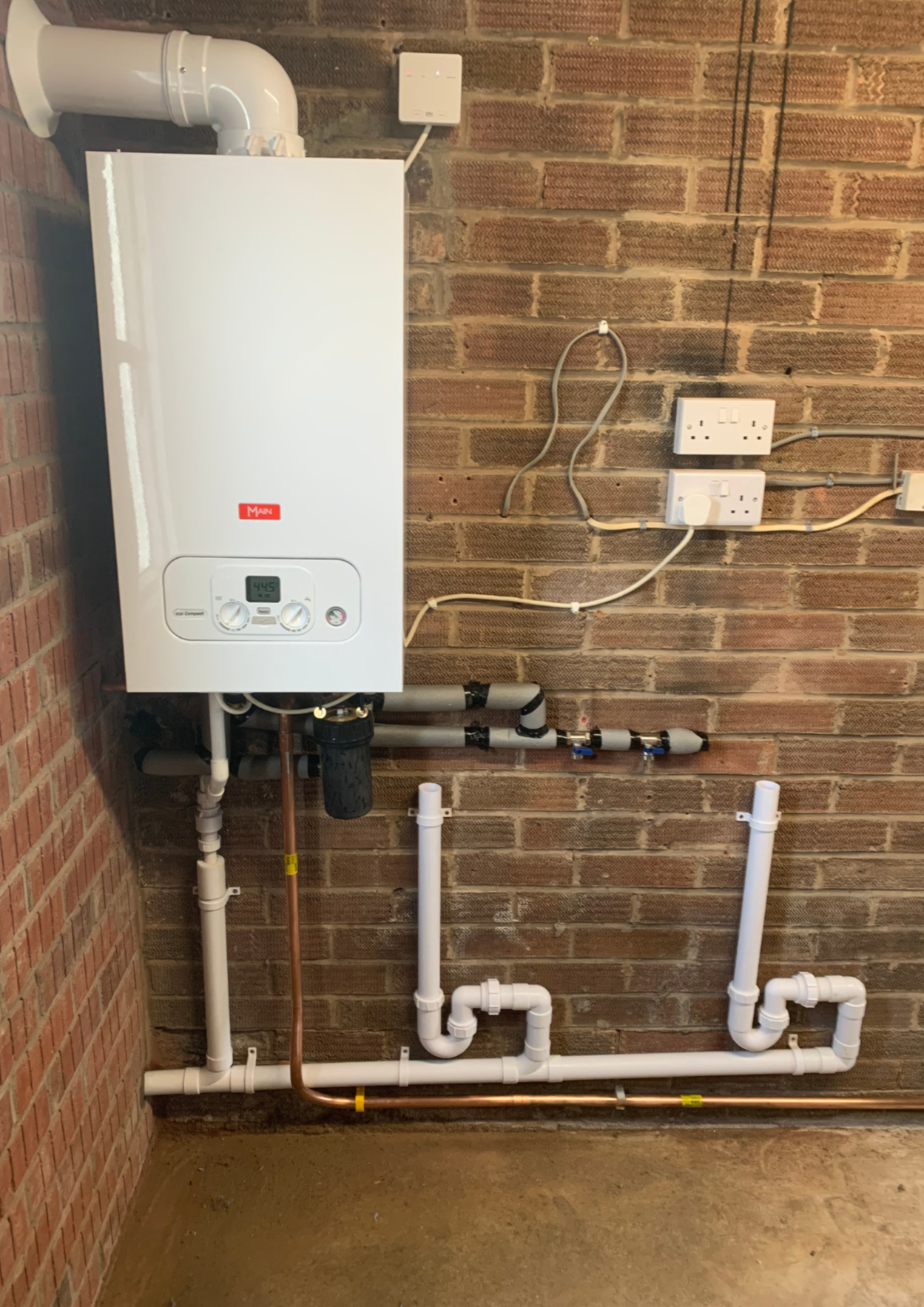 A utility room with a white boiler mounted on a brick wall, connected to PVC and copper pipes, electrical outlets, and a circuit breaker.