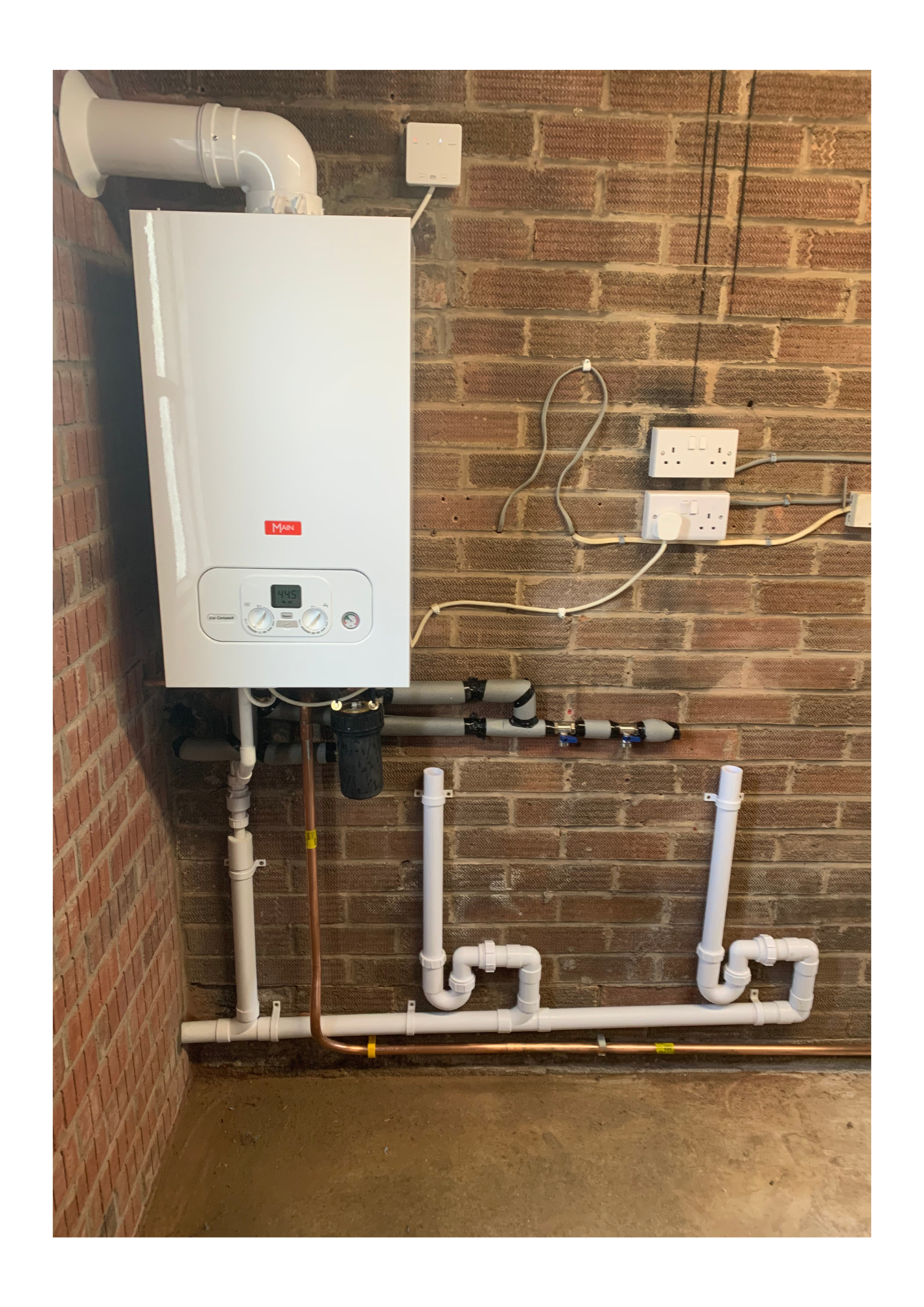 Water heater system with pipes and electrical connections mounted on a brick wall in a utility room.
