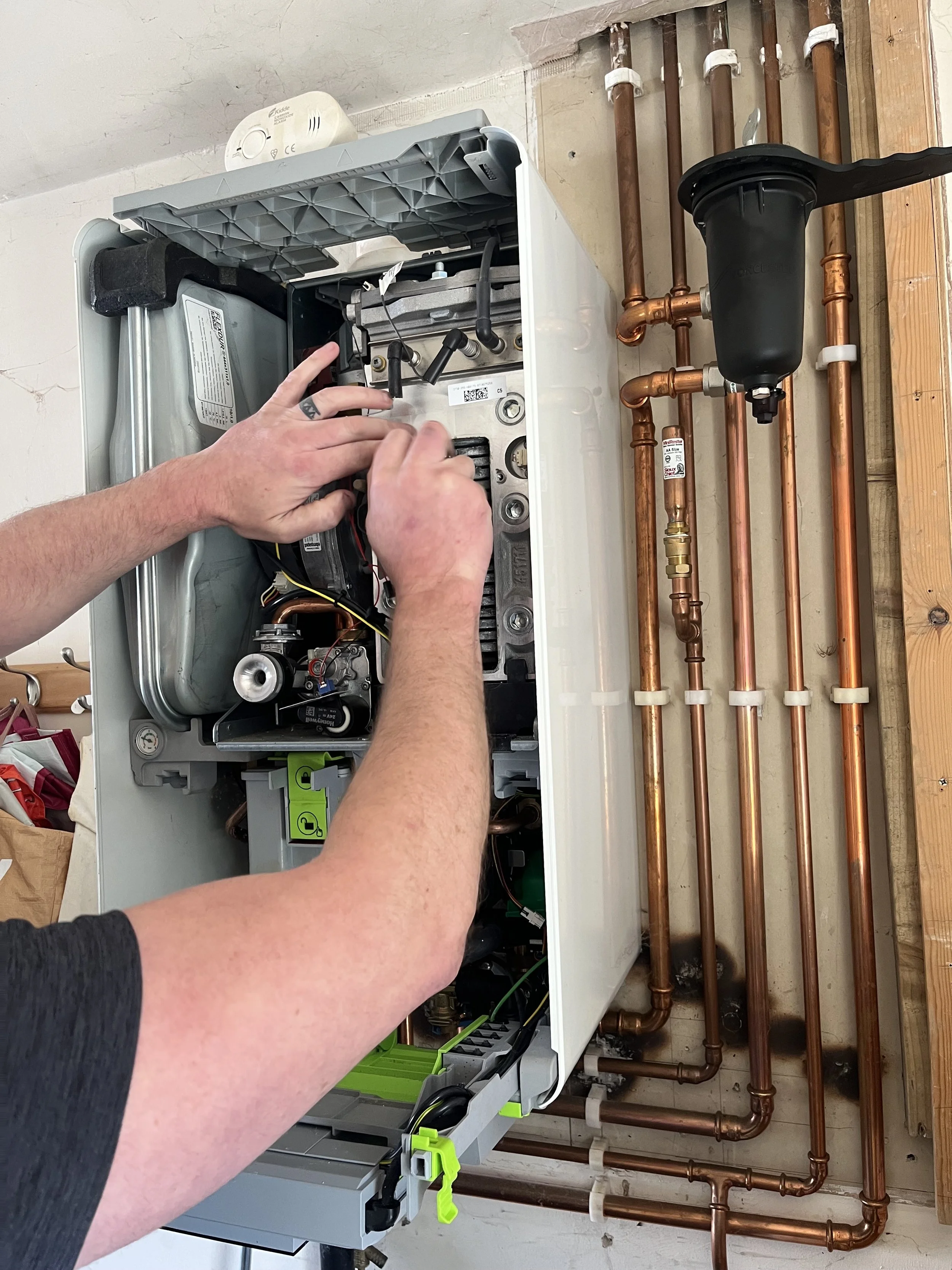 A person working on a heating or plumbing system with exposed copper pipes and a black water filter or pump, inside a building under renovation.