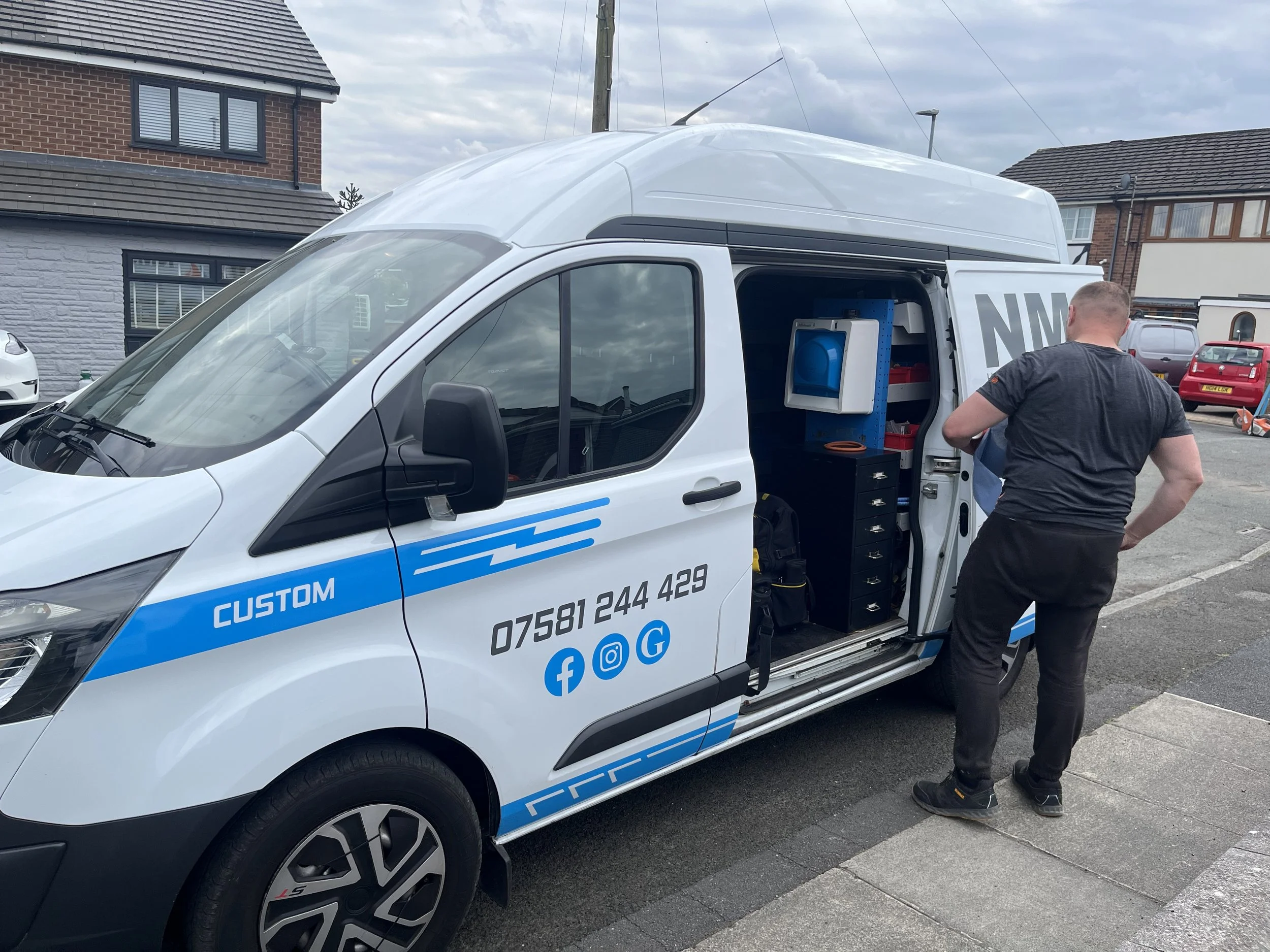 A white service van with blue and black markings parked in a residential area, with a man in black standing beside it. The van's side door is open, revealing storage shelves with tools and equipment. Residential houses and parked cars are visible in the background.
