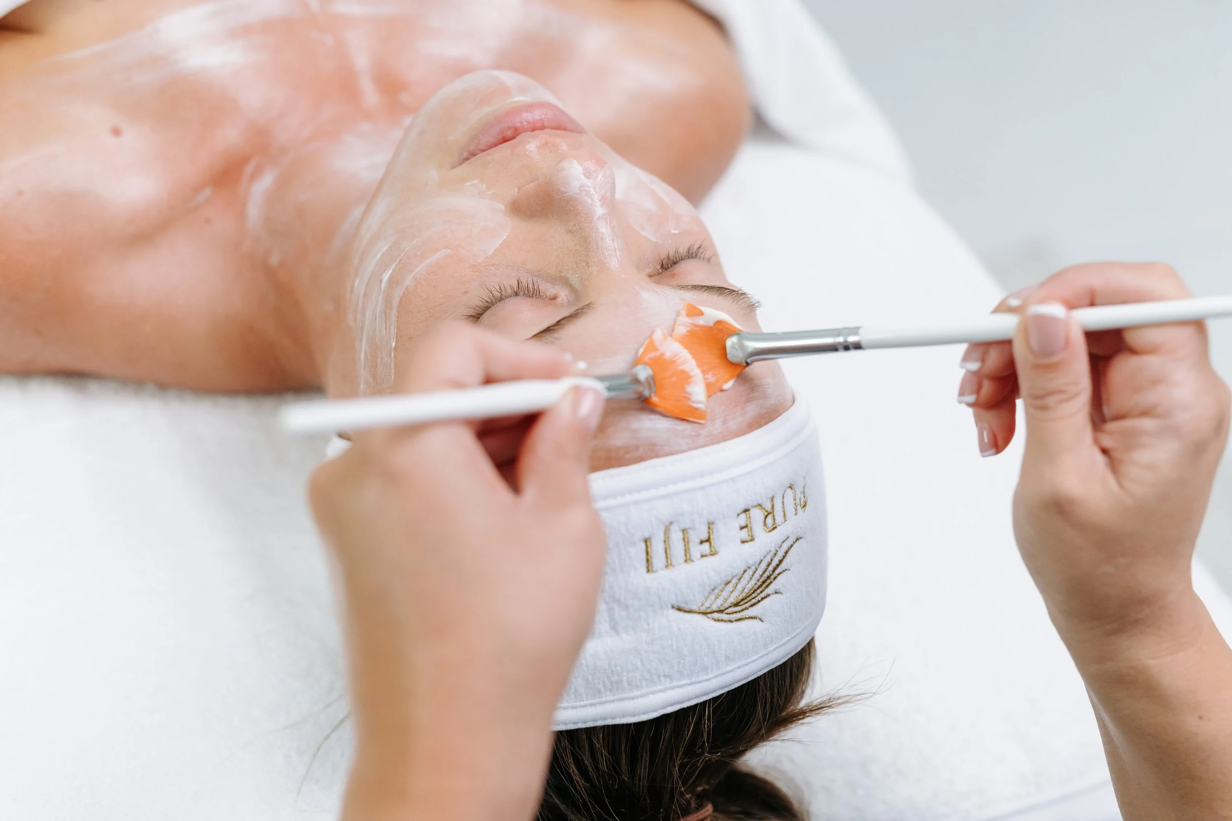 Person receiving a facial massage while lying down with eyes closed, face covered in a white mask.