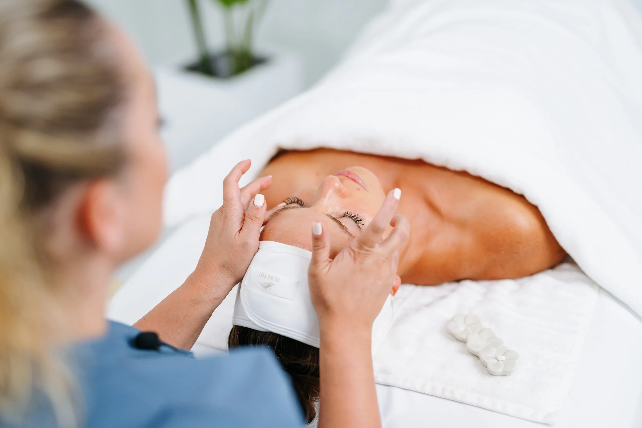 A woman lying on a massage table, receiving a facial treatment from a practitioner. The woman is covered with a white towel and has a facial mask. The practitioner is using her hands to apply or check the facial, and a white headband is holding the woman’s hair back.