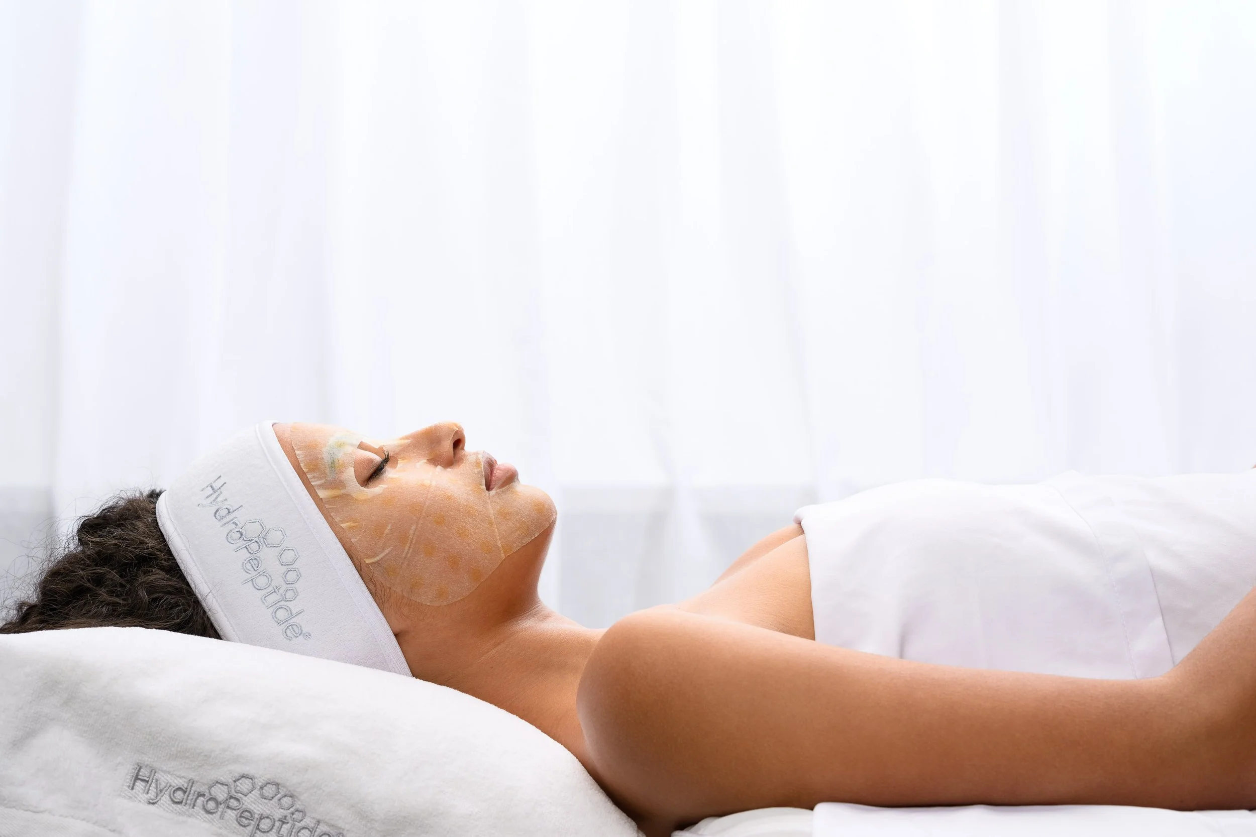 A woman lying on her back with eyes closed in a spa or skincare treatment, wearing a headband and facial mask, in a clean, clinical environment.