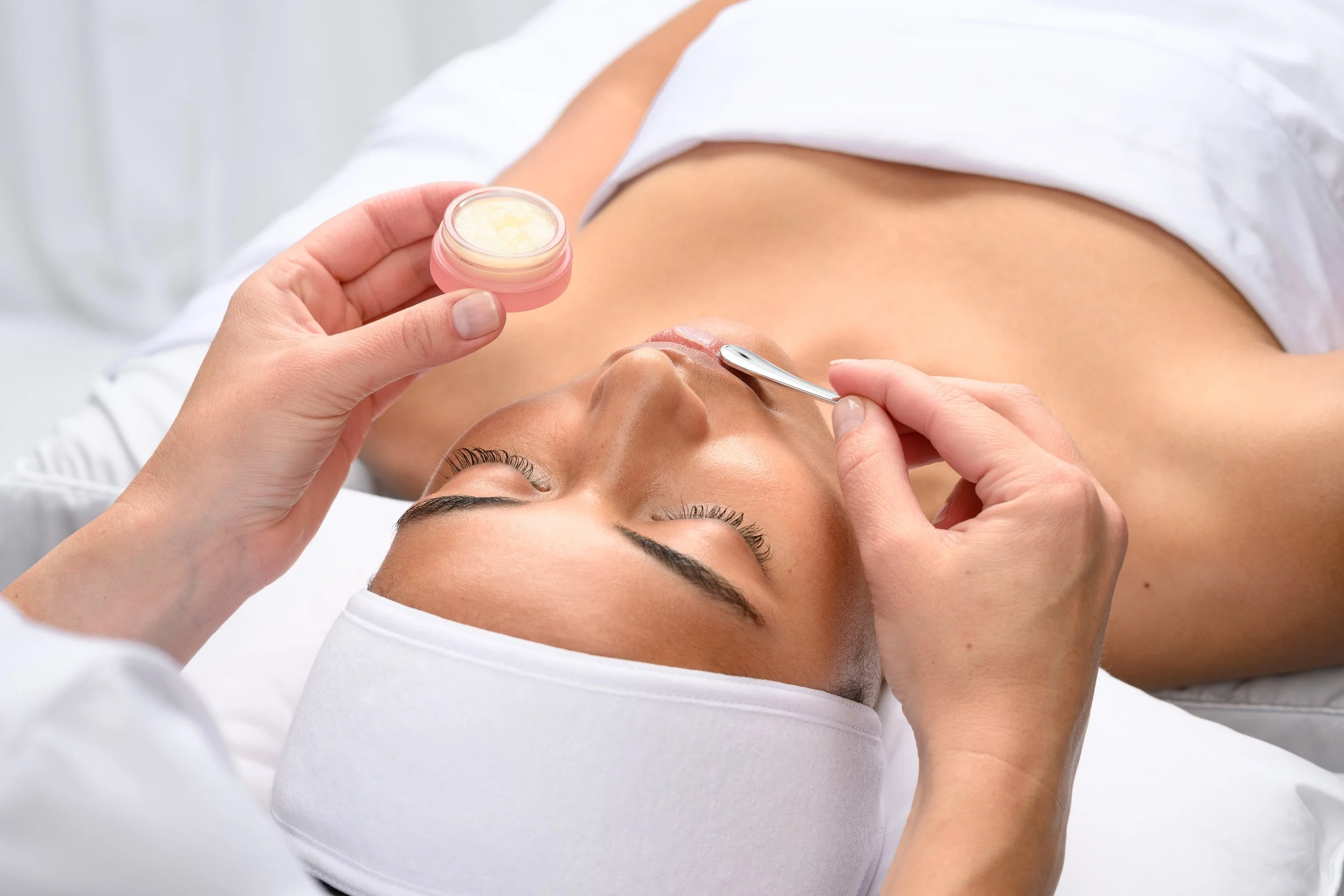 A woman lying down while a person applies lip treatment using a small spatula, holding a pink jar of balm or cream.