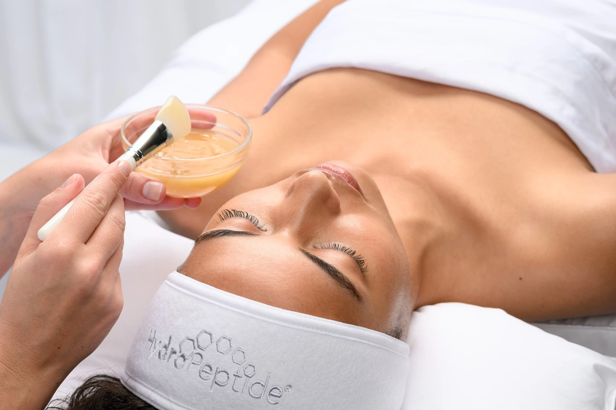 A woman lies on her back receiving a facial treatment with a brush and a bowl of cream or gel.