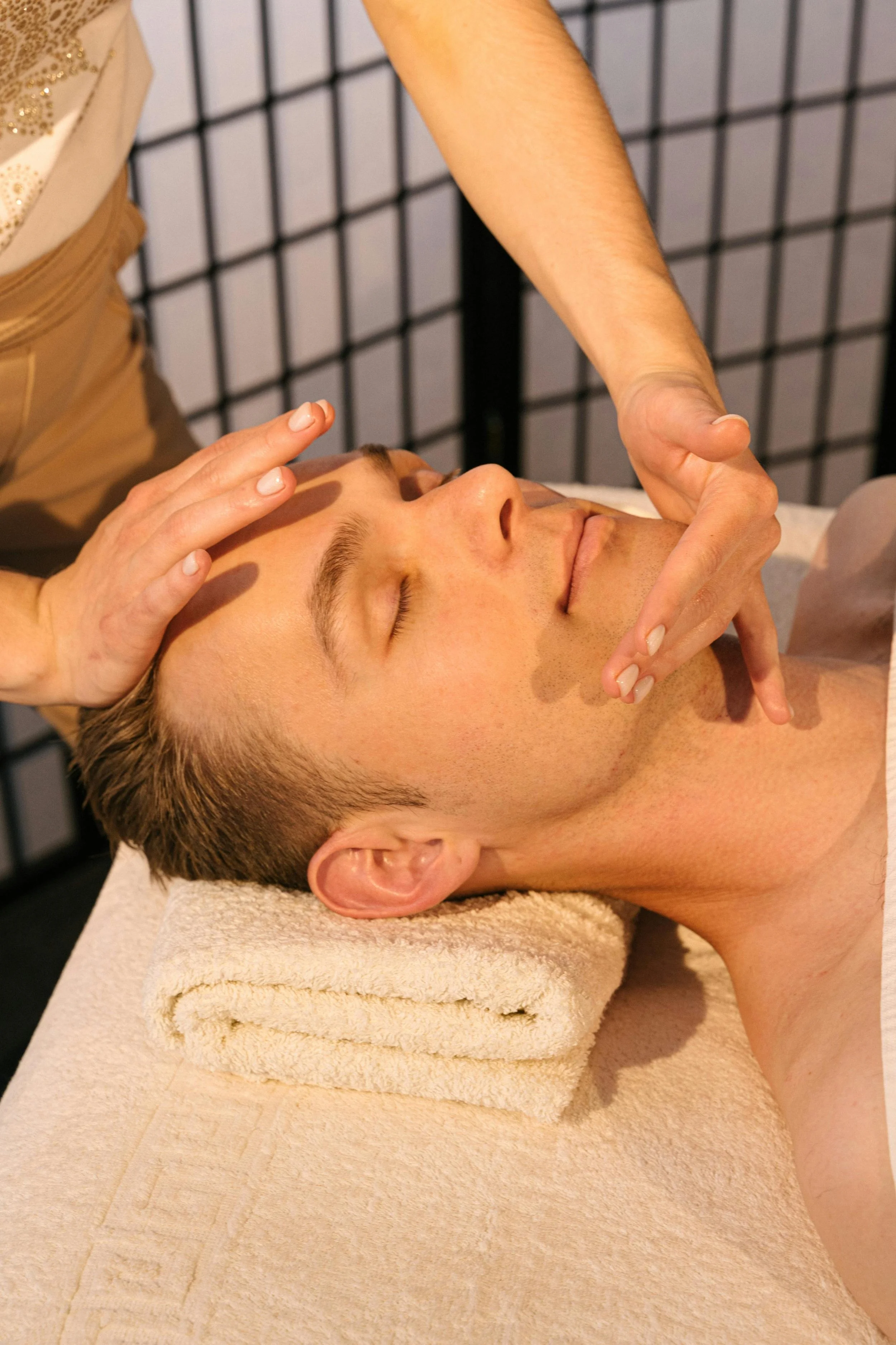 A woman giving a facial massage to a young man lying face up on a massage table. The man has eyes closed and appears relaxed, with a towel under his head. The massage room has a black grid partition in the background.