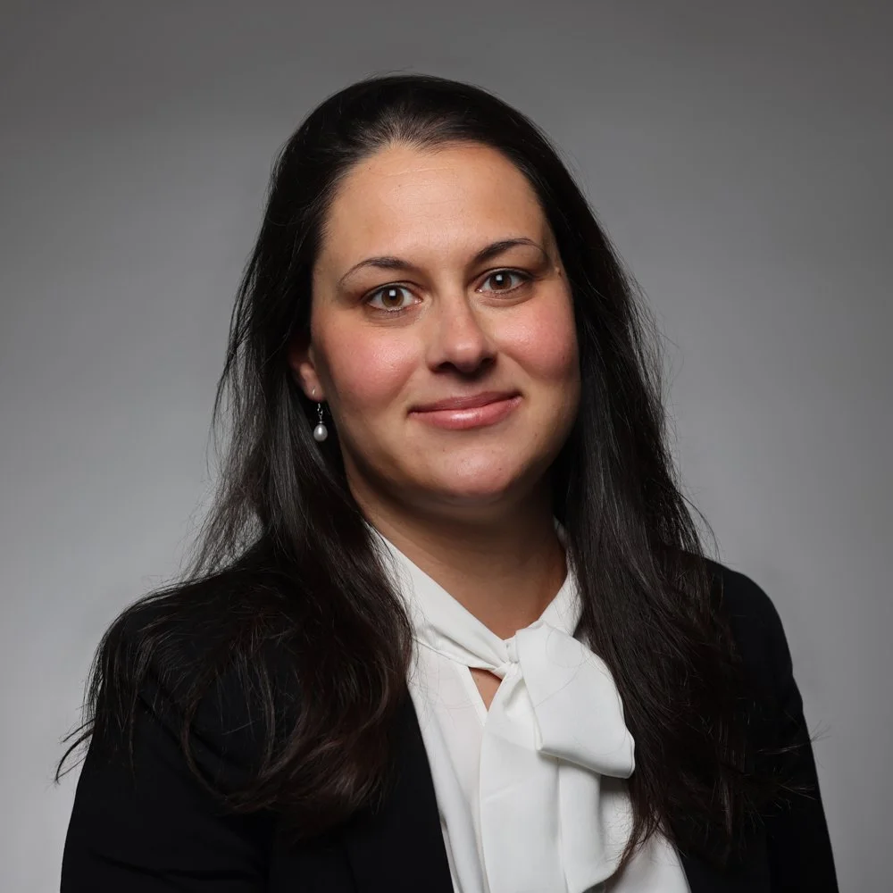 Portrait d'une femme portant un chemisier blanc avec un nœud et un blazer noir contre un fond gris.