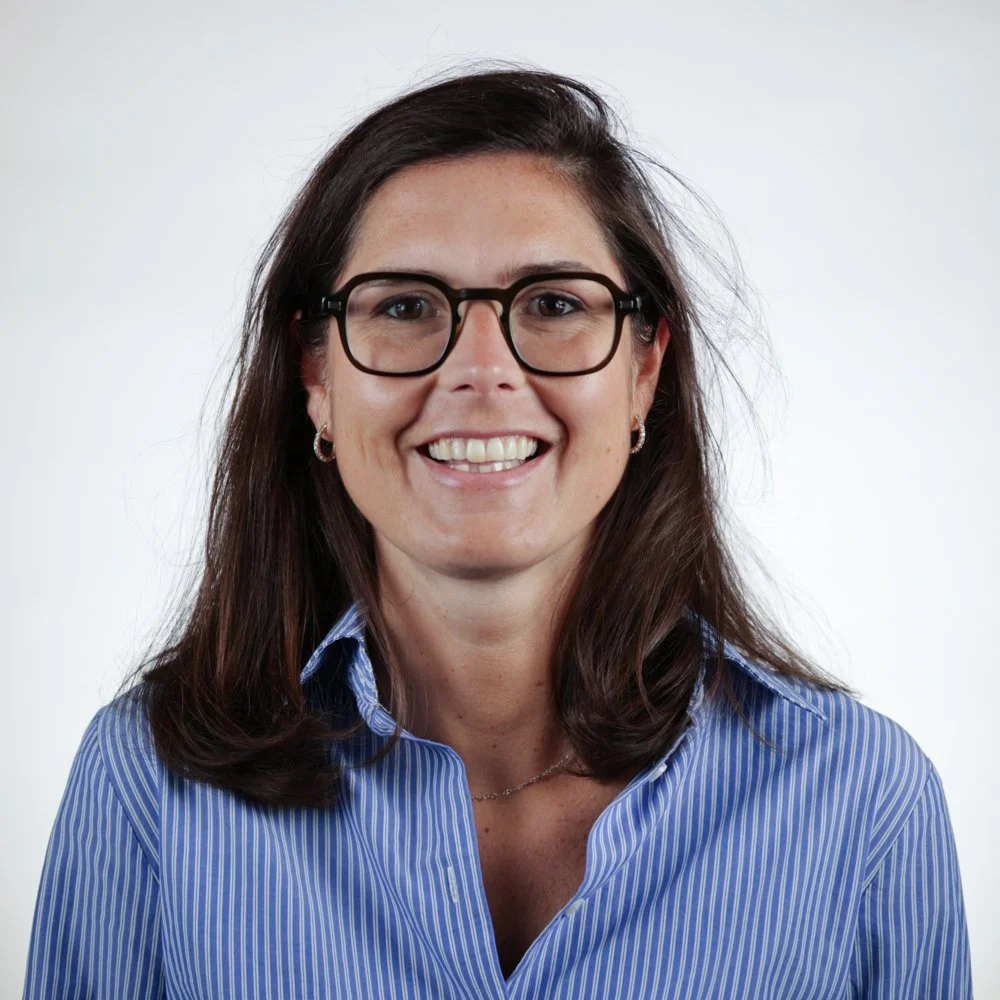 Une femme avec des cheveux bruns, portant des lunettes noires et un chemisier à rayures bleues, sourit devant un fond blanc.