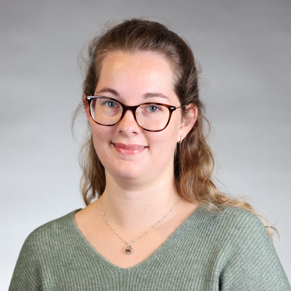 Une femme porte des lunettes marron, un collier et un pull vert clair, avec un sourire léger, contre un fond gris clair.