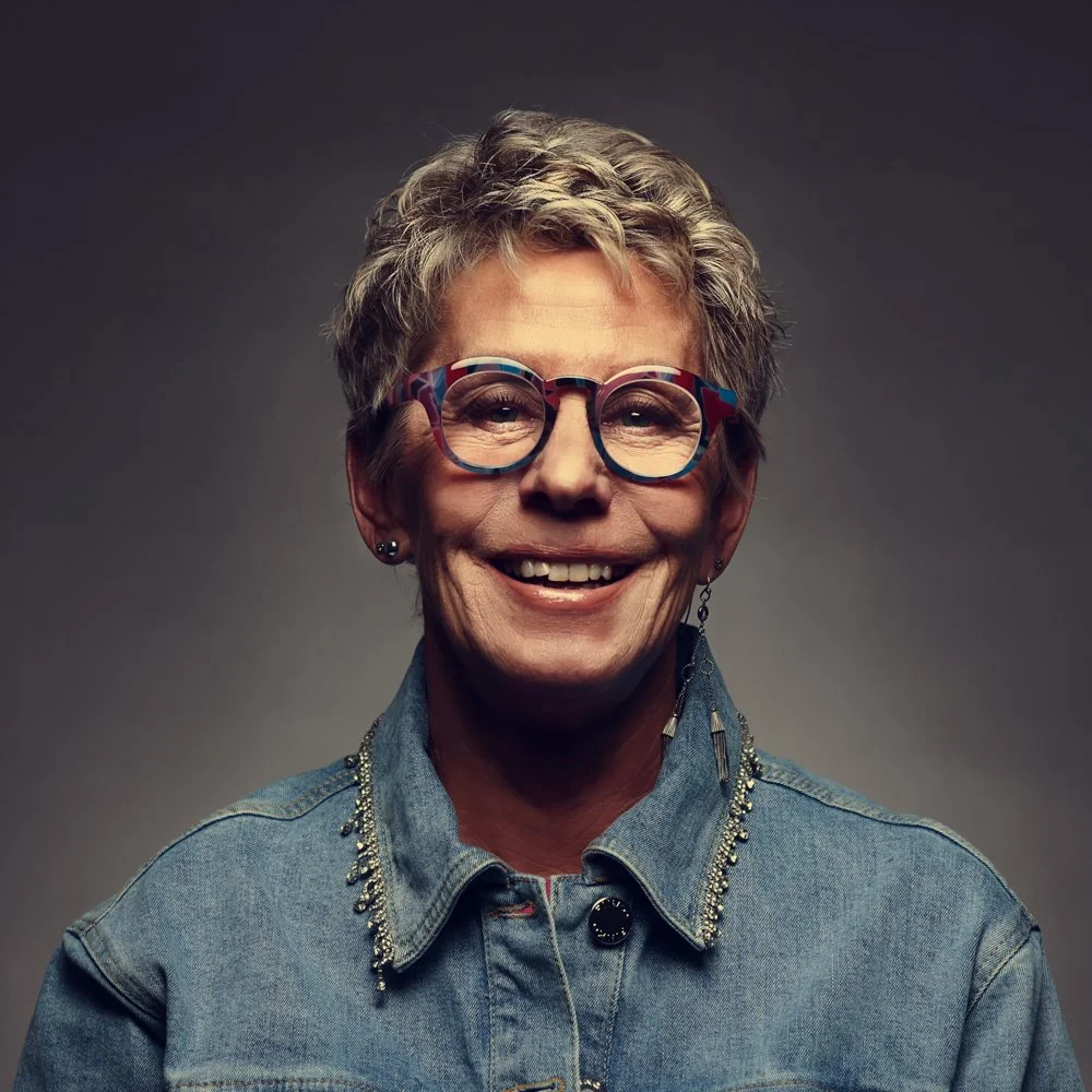 Portrait d'une femme âgée souriante portant des lunettes colorées, une veste en jean avec des détails scintillants, et des boucles d'oreilles, face à un arrière-plan gris.