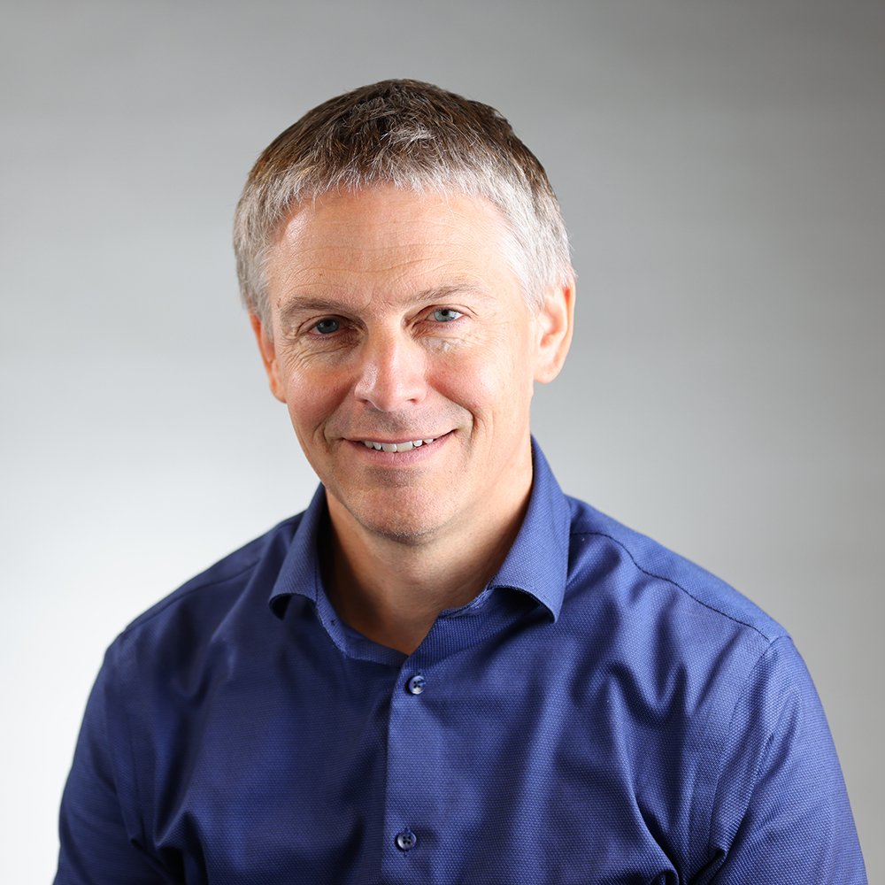 Un homme souriant avec des cheveux courts et gris, portant une chemise bleue, portrait en gros plan contre un fond neutre.