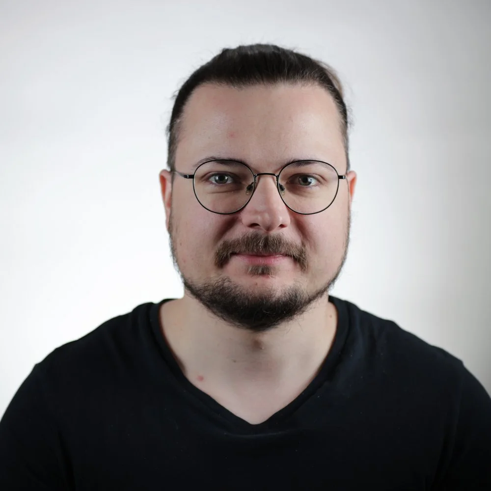 Portrait d'un homme portant des lunettes rondes, avec une barbe et une moustache, portant un t-shirt noir, contre un fond blanc.
