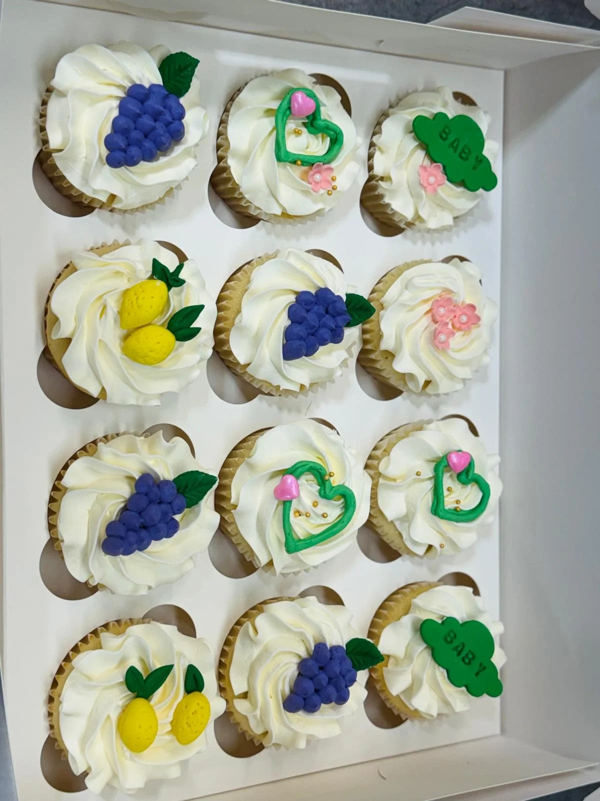 A tray of decorated cupcakes with white icing and fruity-themed toppers, including grapes, lemons, and hearts, some with the word 'BABY' written on green cloud-shaped toppers.