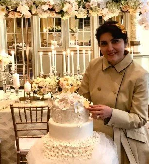 Woman in beige coat standing next to a wedding cake decorated with flowers, in a decorated event space with floral arrangements and candlelights.