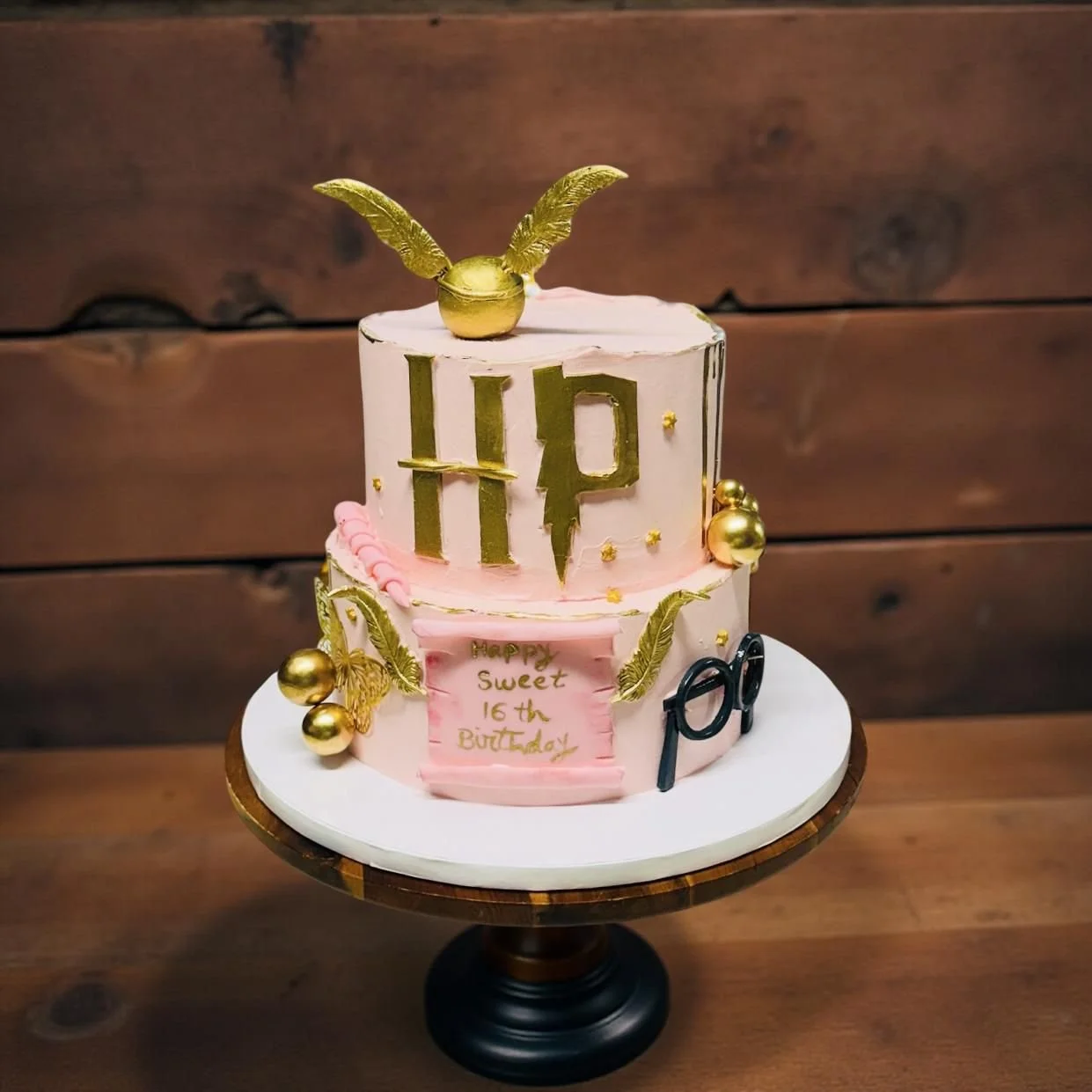 A two-tier birthday cake with pink and white frosting, decorated with gold leaves and balls. The top tier features a gold Harry Potter lightning bolt and glasses. The cake has a pink plaque with gold writing that says, "HAPPY SWEET 16TH BIRTHDAY," an