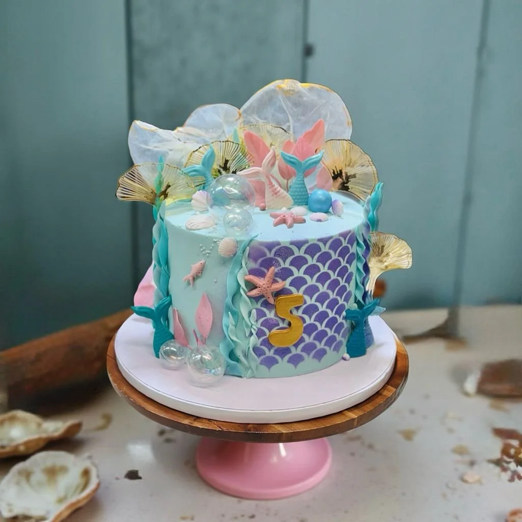A colorful underwater-themed birthday cake decorated with mermaid tails, shells, starfish, and seaweed, featuring a purple scale pattern, a gold number 5, and blue, pink, and turquoise accents.
