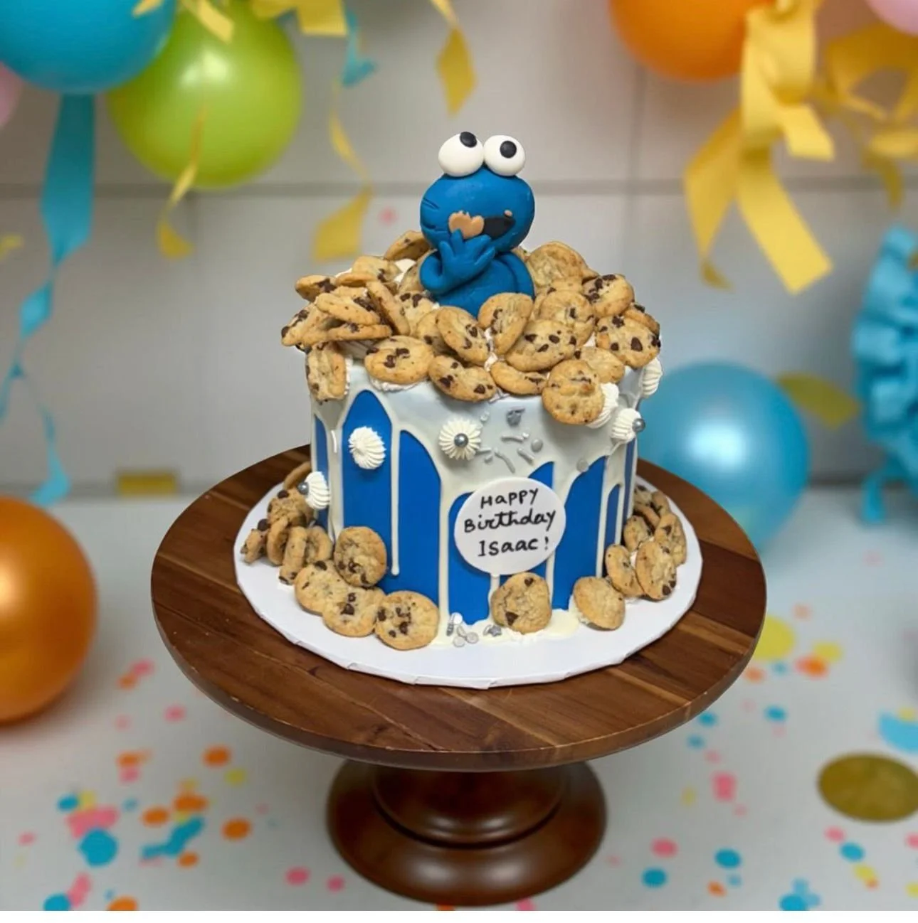 A birthday cake decorated with chocolate chip cookies, a blue cookie monster figure on top, and a note that says "Happy Birthday Isaac!" It is set on a wooden cake stand with colorful balloons and confetti in the background.