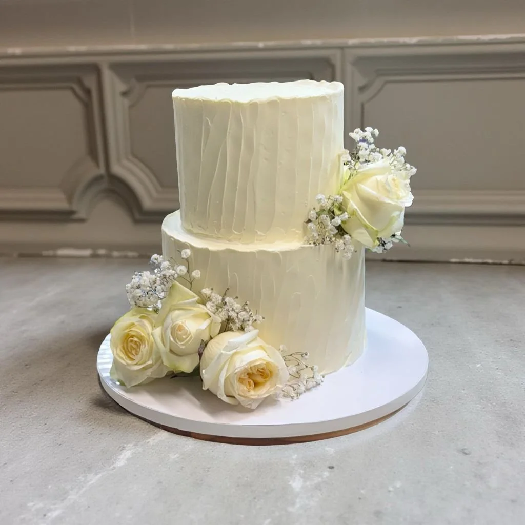 A two-tier white wedding cake decorated with white roses and baby's breath flowers, placed on a white circular cake board.