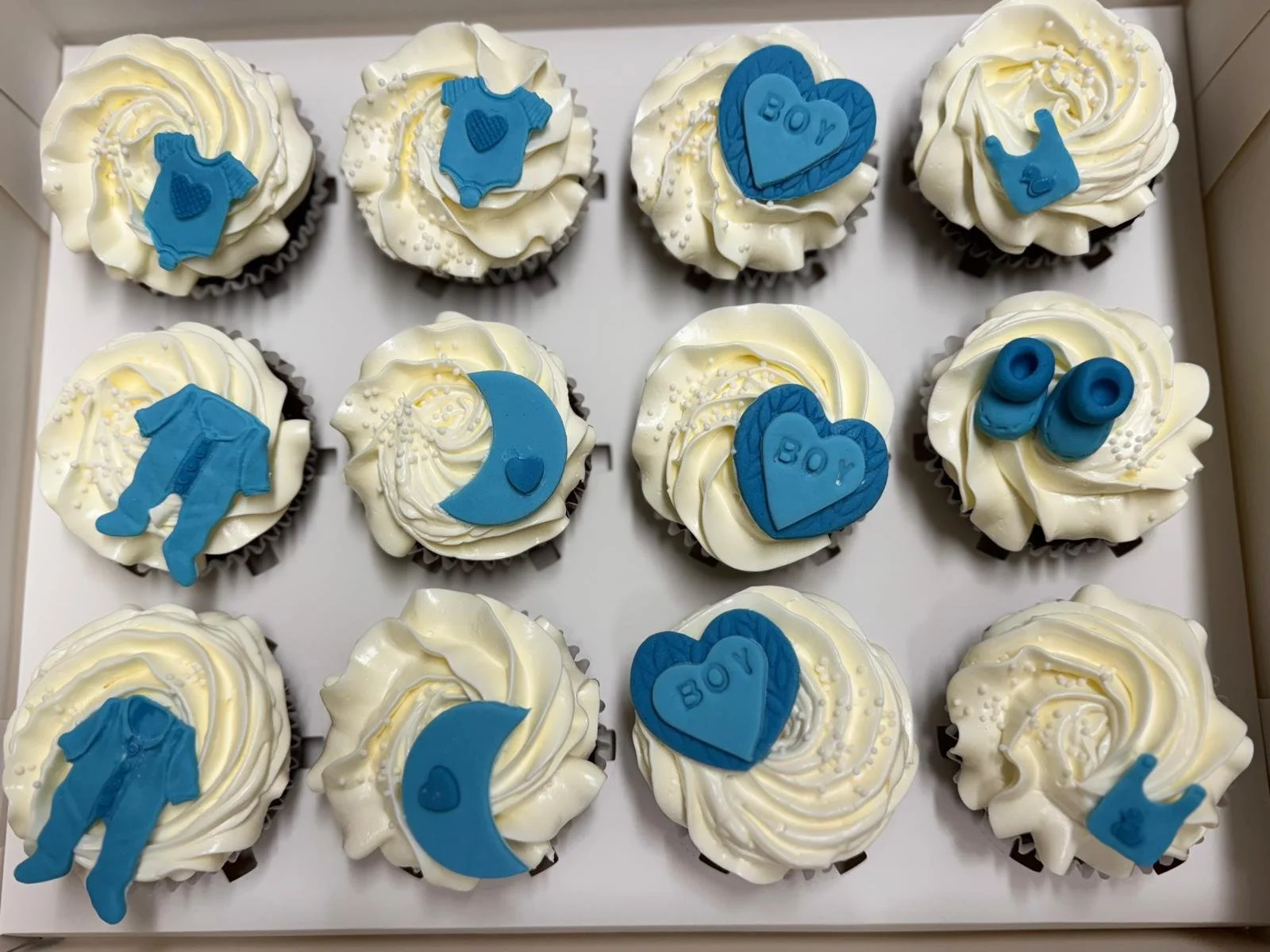 A box of 12 cupcakes decorated with white frosting and blue themed toppers, including baby onesies, hearts, moons, and the word 'BOY'.