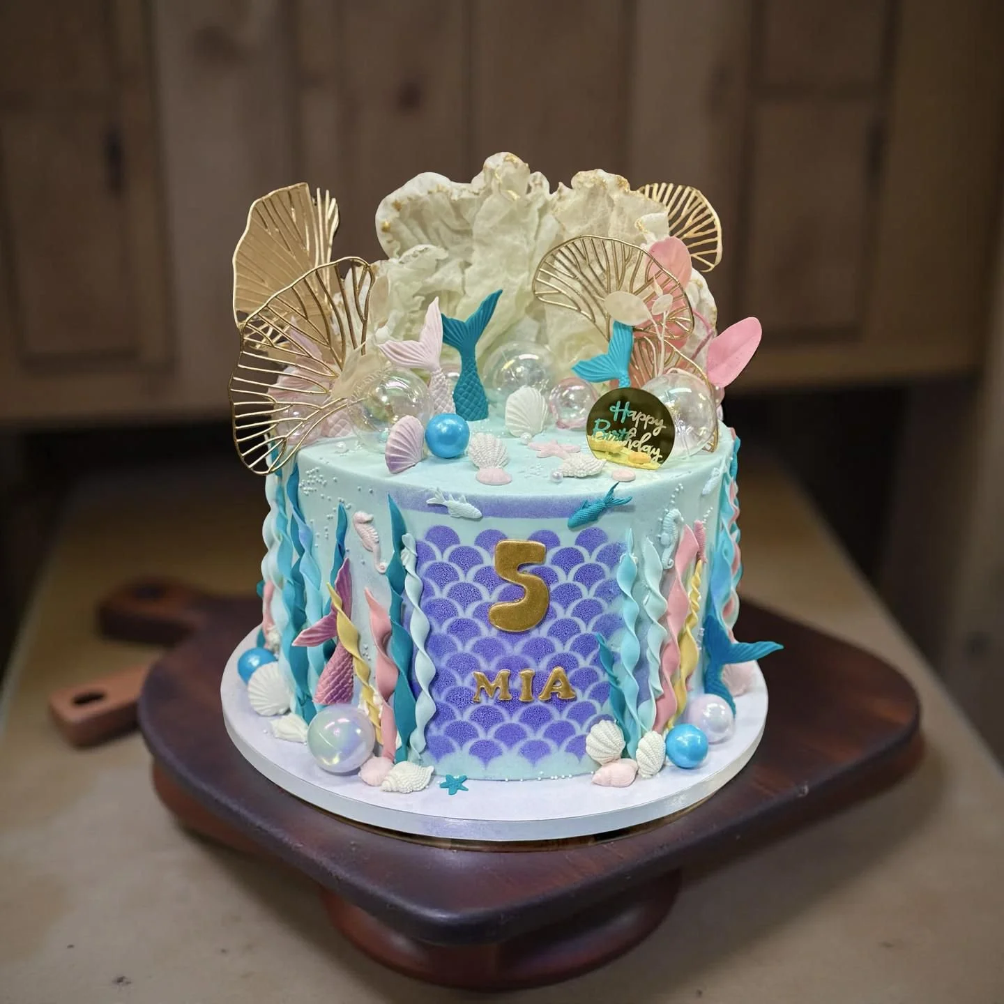 A birthday cake decorated with mermaid and ocean-themed decorations, including shells, fish, and mermaid tails, with a gold number 5 and the name Mia, on a wooden cake stand.