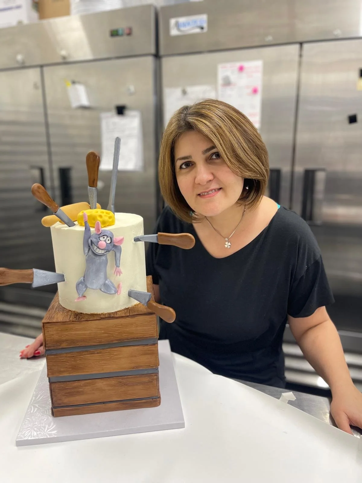 Woman with short brown hair smiling next to a cake decorated with a mouse figure and kunai knives sticking out, in a kitchen setting.