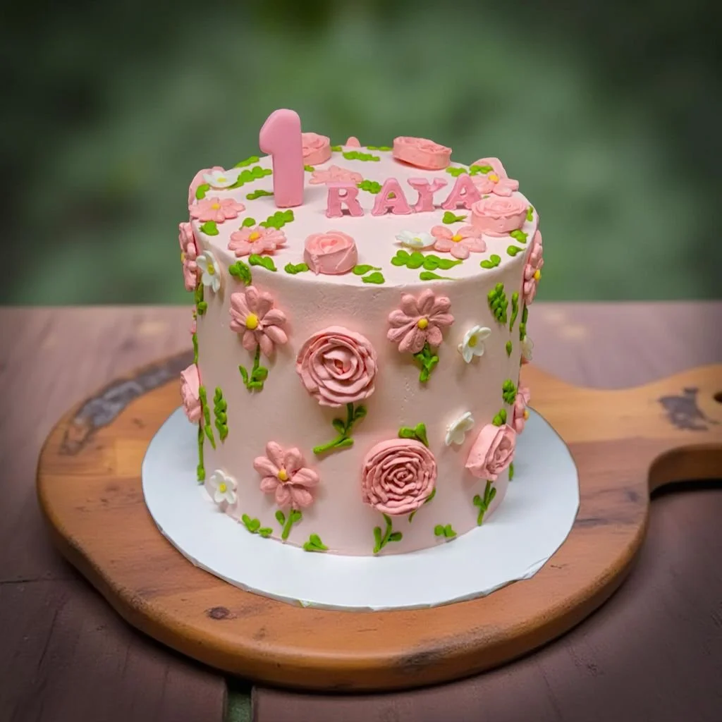 A pink birthday cake decorated with pink and white flowers, green leaves, and a pink number one candle, with the name 'Raya' on top, placed on a wooden cutting board.