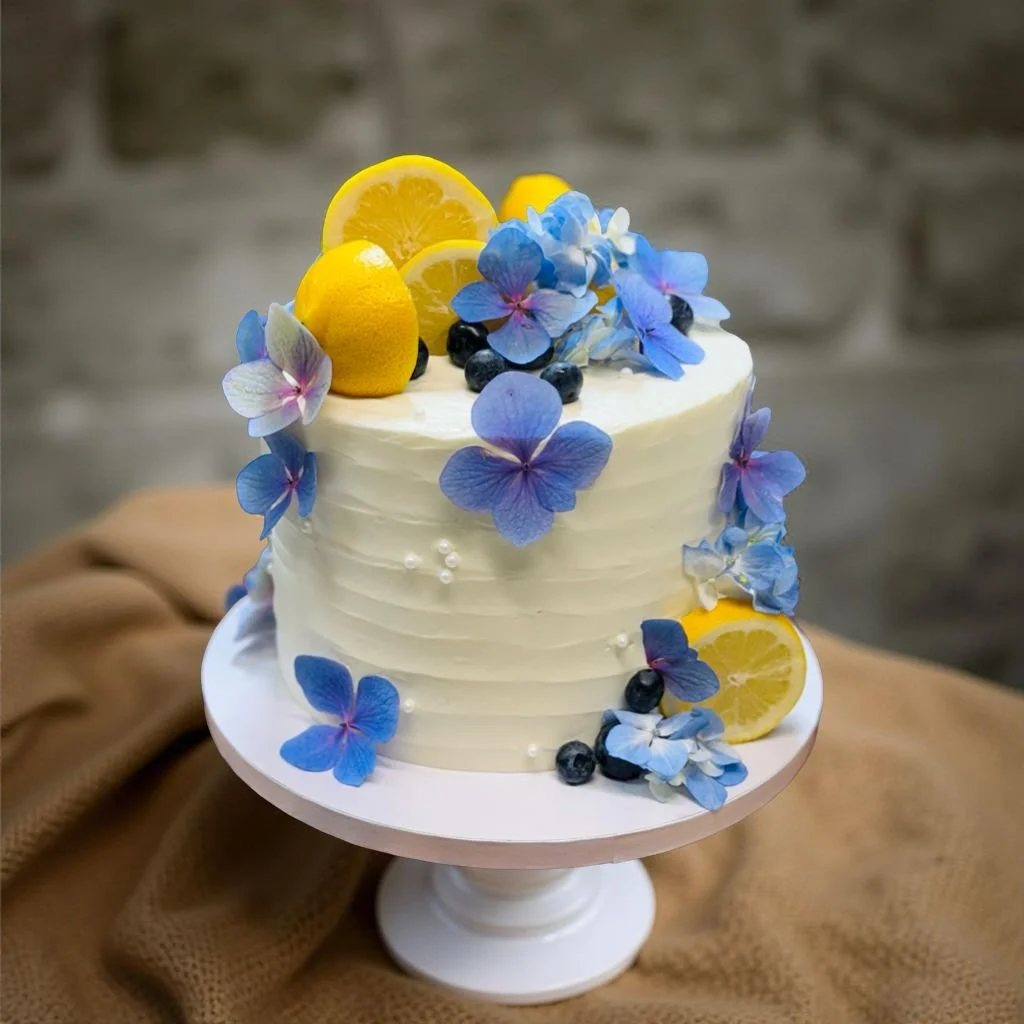 A white frosted cake decorated with blue and purple hydrangea flowers, lemon slices, blueberries, and small white edible pearls, displayed on a white cake stand.
