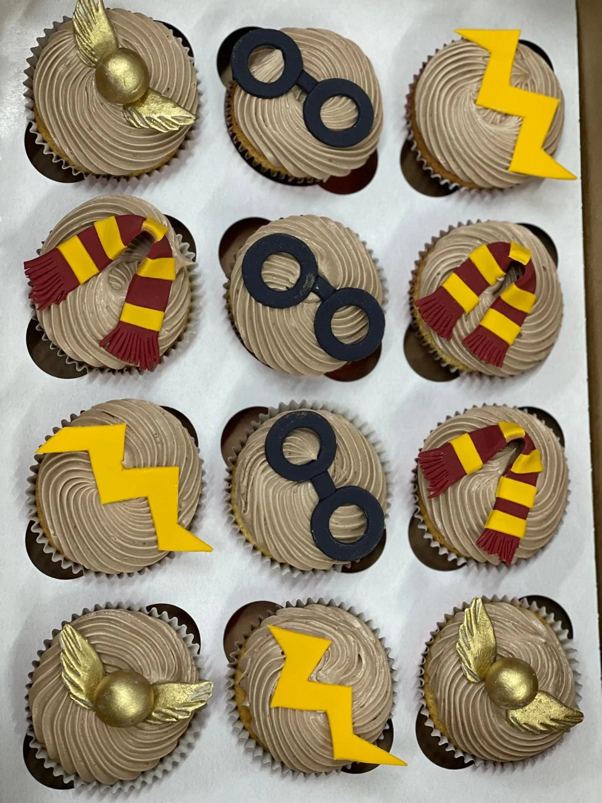 Box of cupcakes decorated with Harry Potter theme. Some cupcakes have gold wings and a gold sphere, others have black glasses, yellow and red lightning bolts, and yellow and red scarves.