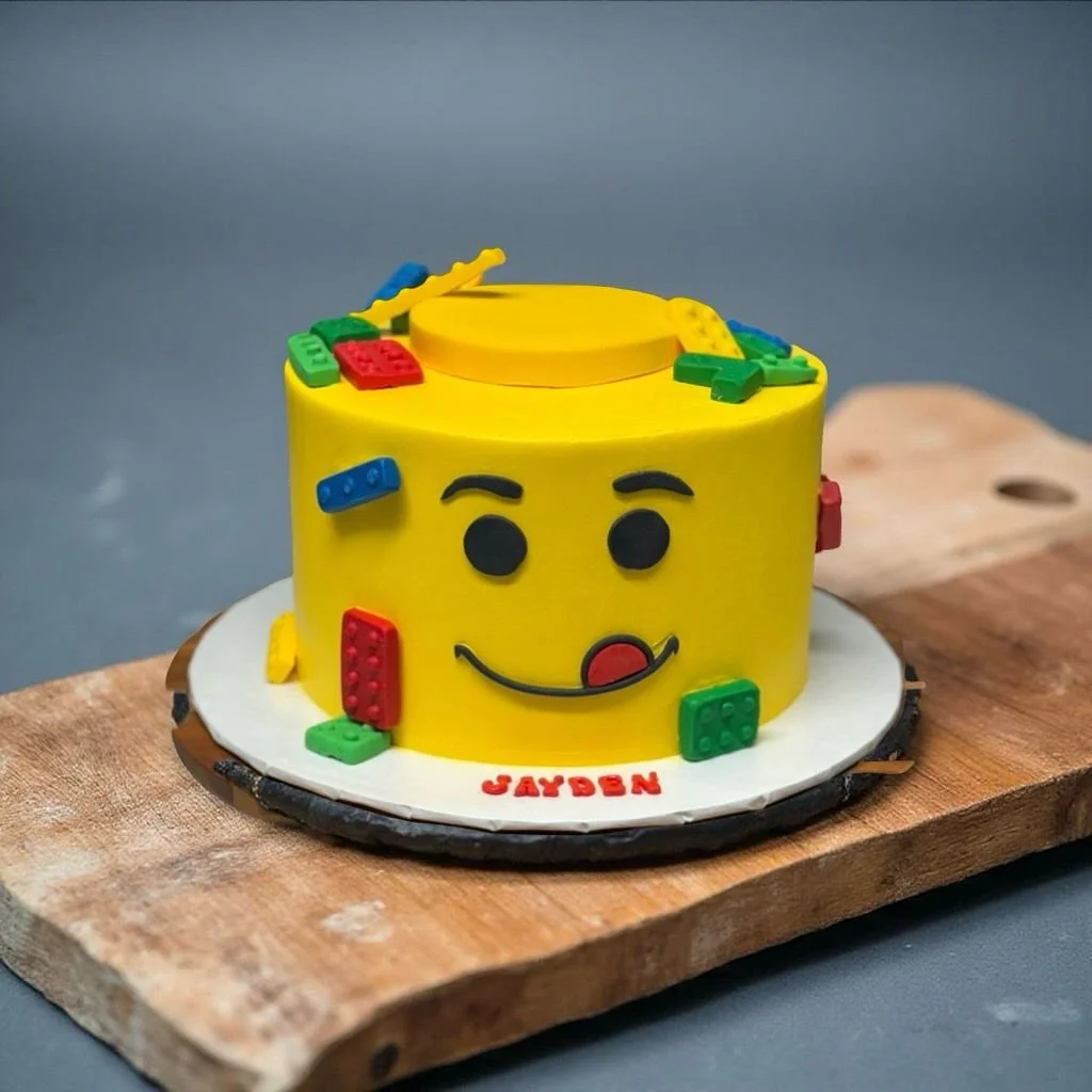 Lego-themed birthday cake with a yellow face, black eyes, eyebrows, and a red tongue, decorated with colorful Lego pieces, on a wooden board.