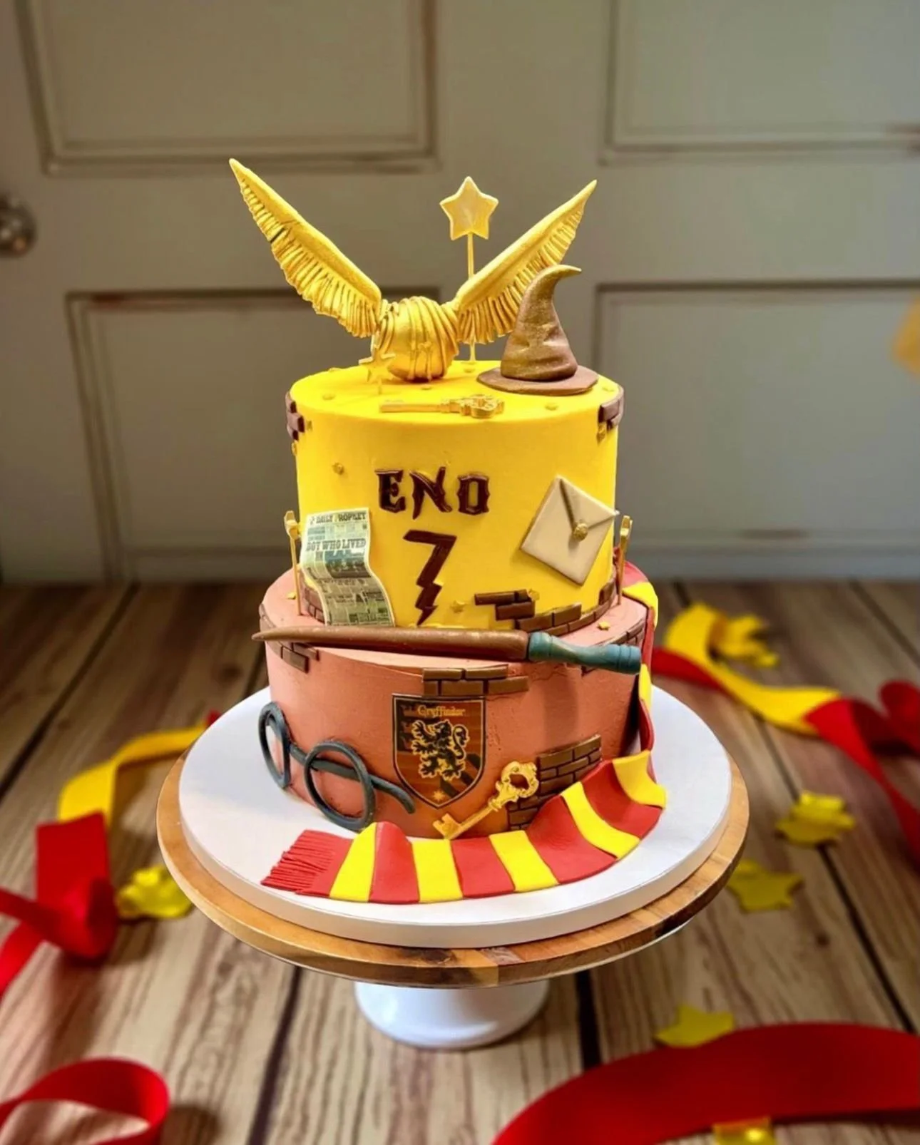 A two-tier cake decorated with Harry Potter-themed fondant and decorations, including a yellow winged sculpture with a star, a witch's hat, glasses, a wand, a shield with a lion, and the word "END" with the number "7" on the upper tier, and striped r